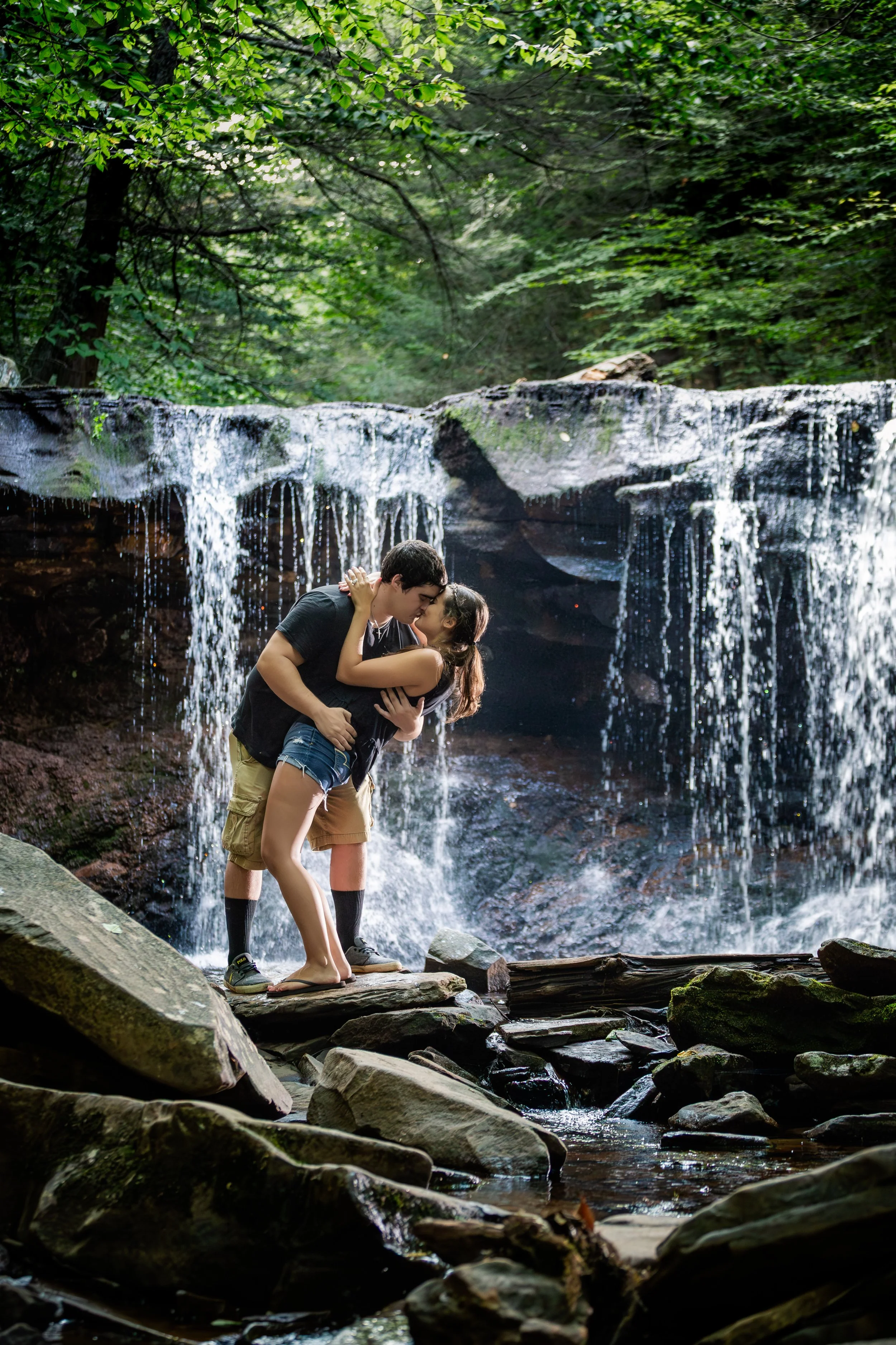A young couple sharing a kiss in front of a small waterfall in a lush forest, standing on rocks.