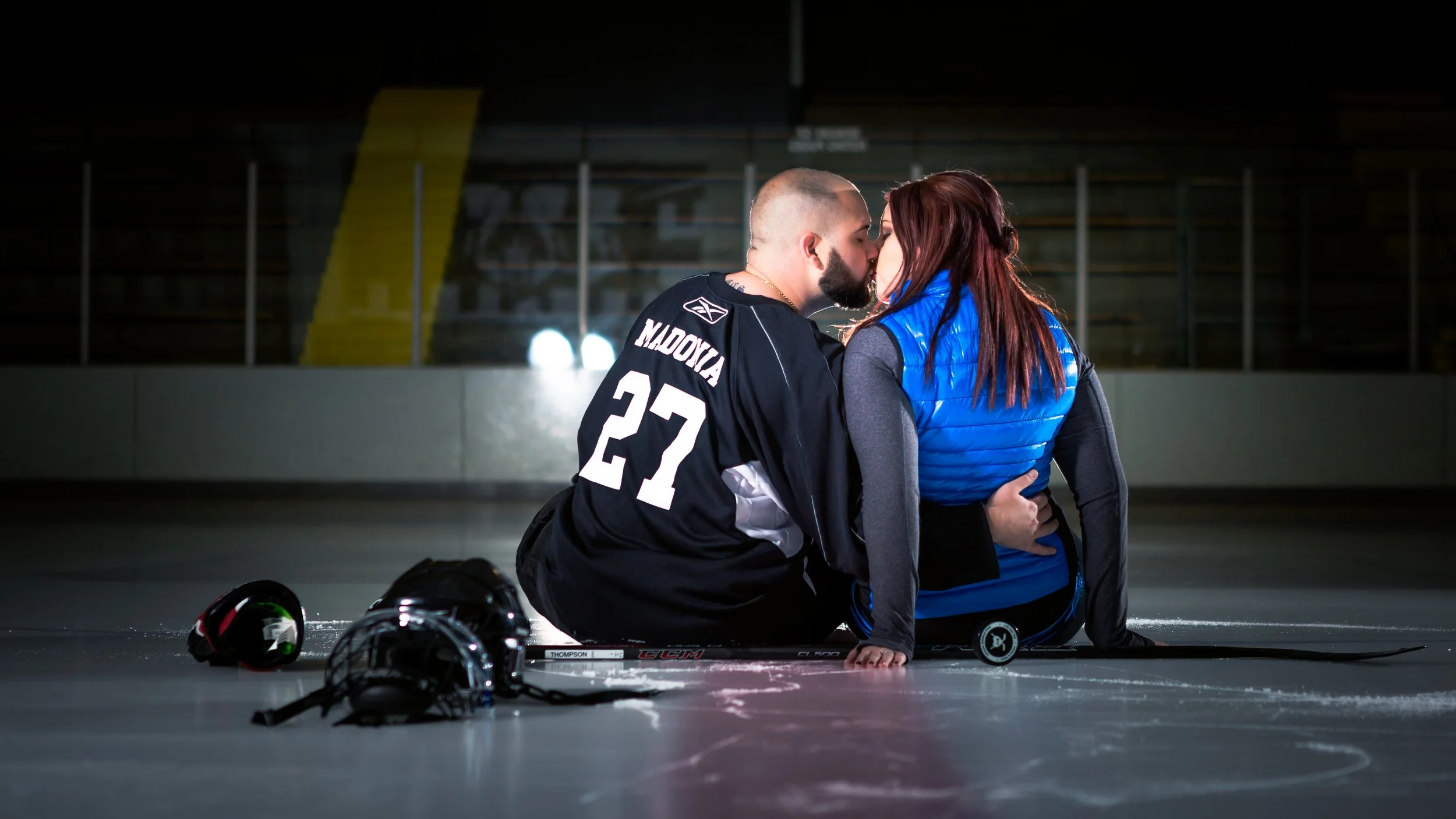 Hockey player and woman kissing on an indoor ice rink, helmets and hockey sticks nearby.