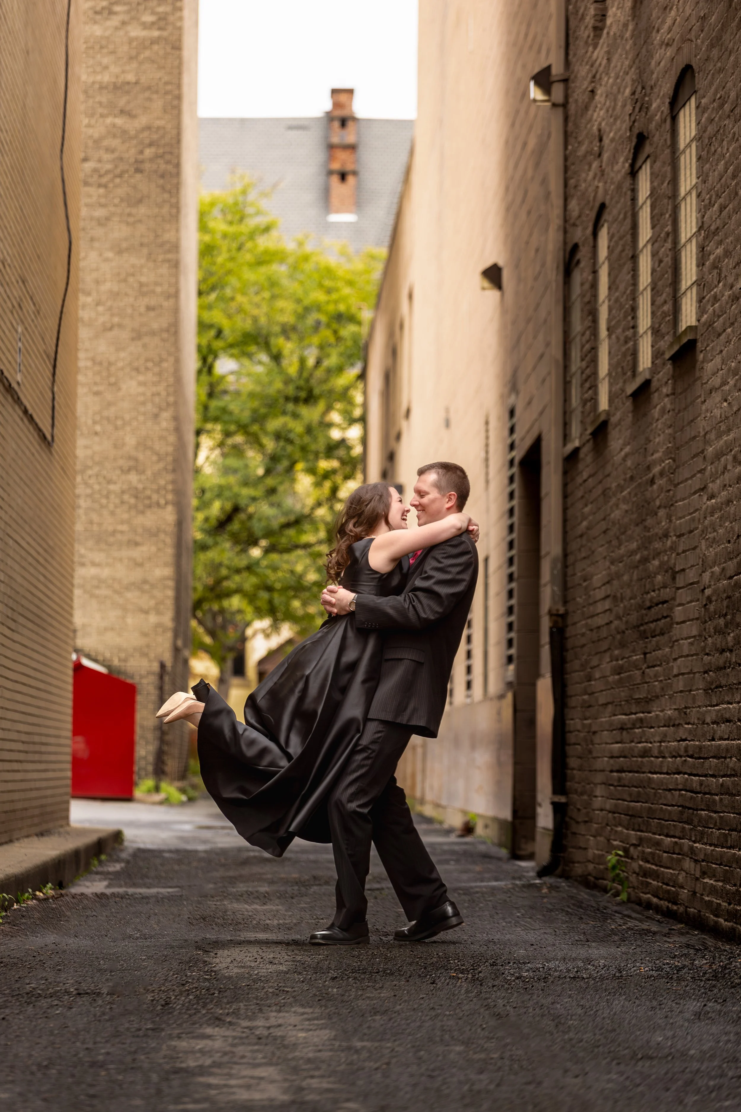 A happy couple dressed in formal attire embracing and dancing in a narrow urban alleyway with brick walls on each side, with trees and buildings visible in the background.