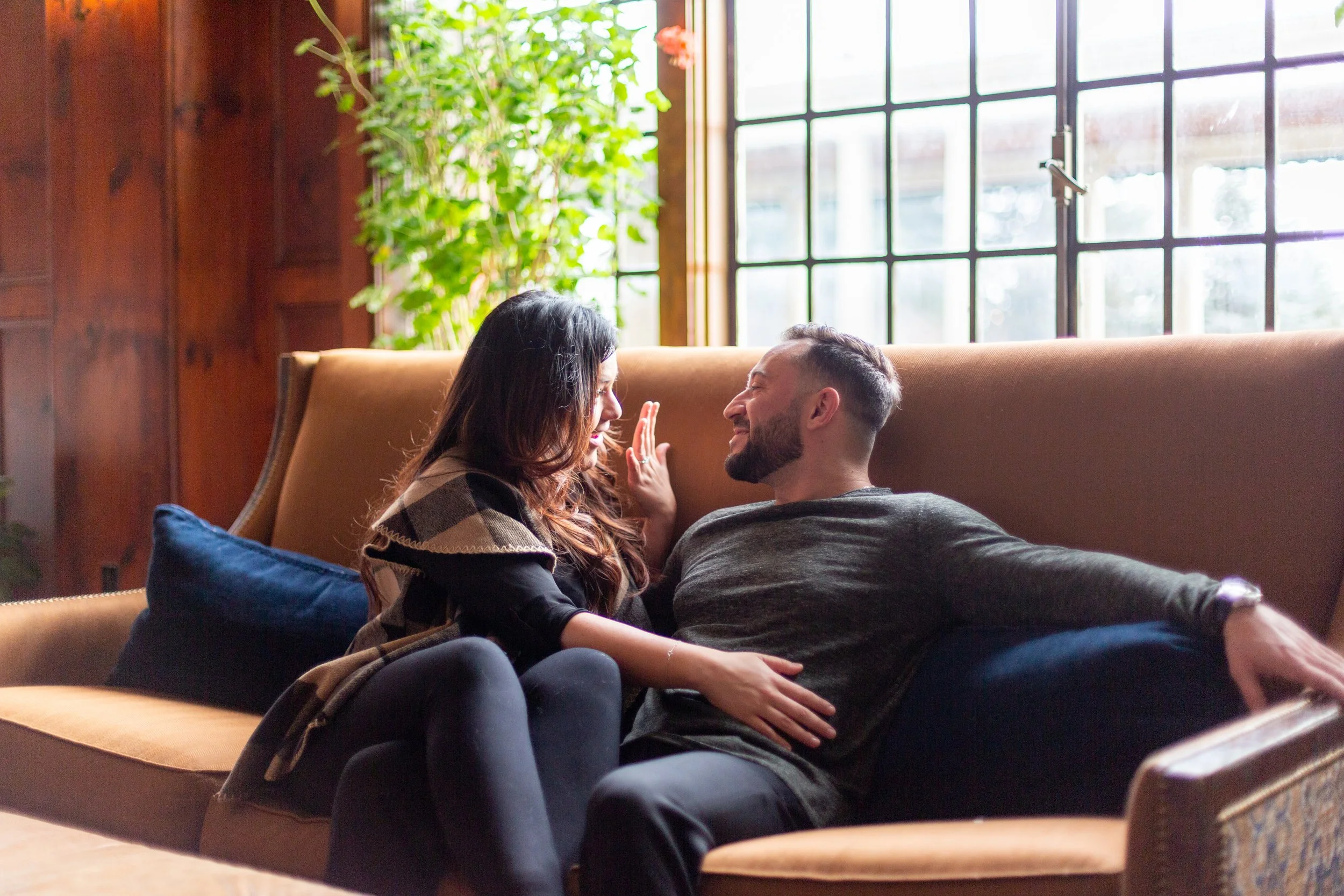 A couple lounging on a tan sofa near large windows, smiling and engaging with each other in a cozy, well-lit room.