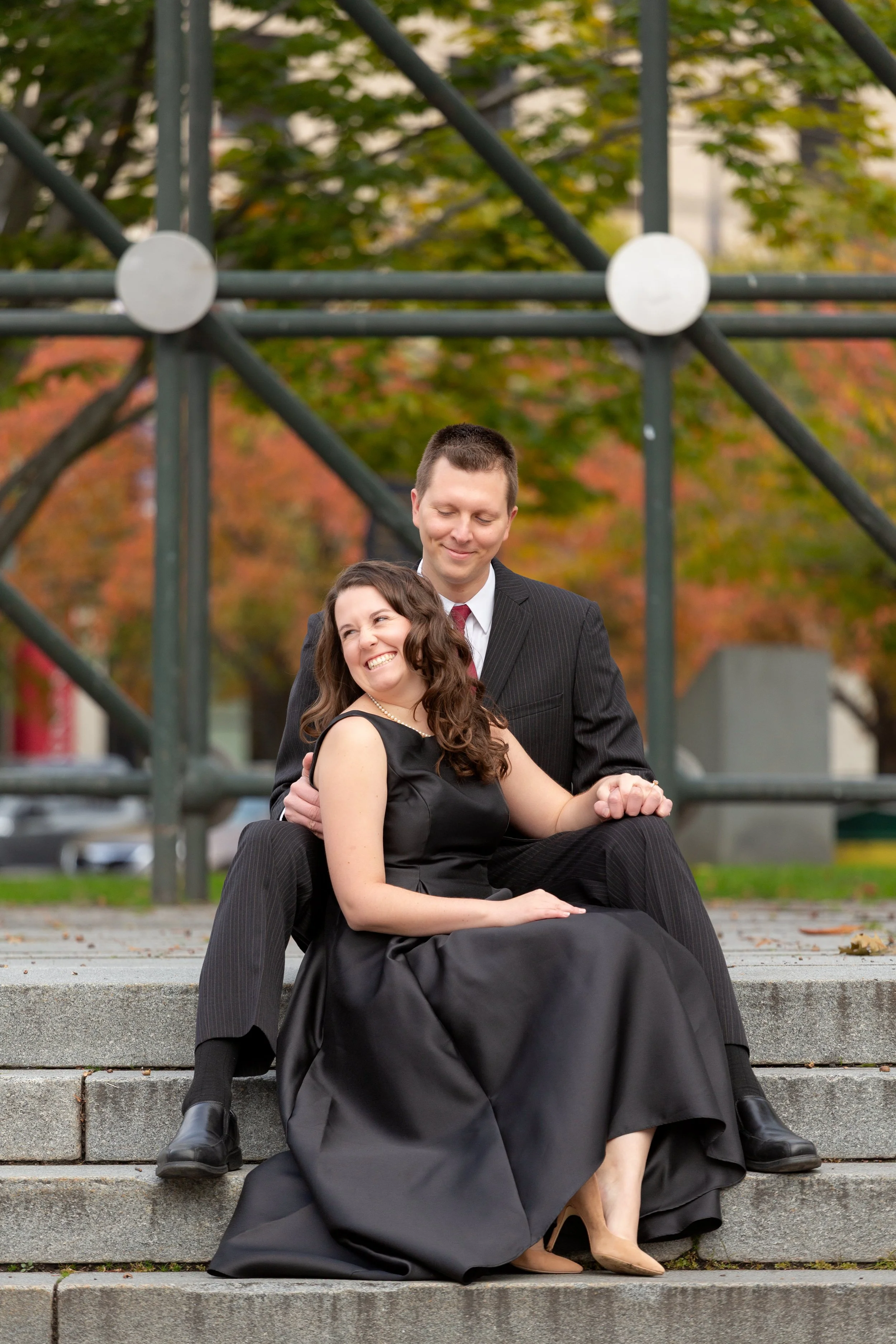 A smiling woman in a black dress and beige heels sitting on steps with a man in a black suit behind her, both holding hands and enjoying time outdoors with colorful autumn trees and metal playground equipment in the background.