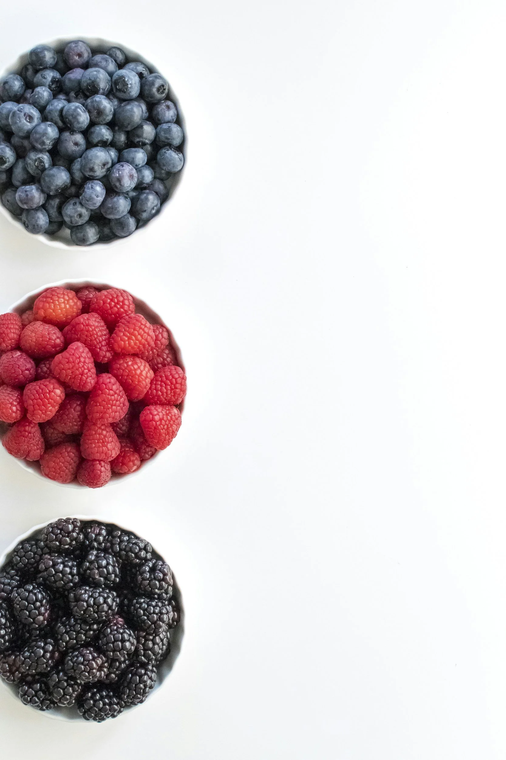 Three small bowls of berries, from top to bottom: blueberries, raspberries, blackberries, on a white surface.