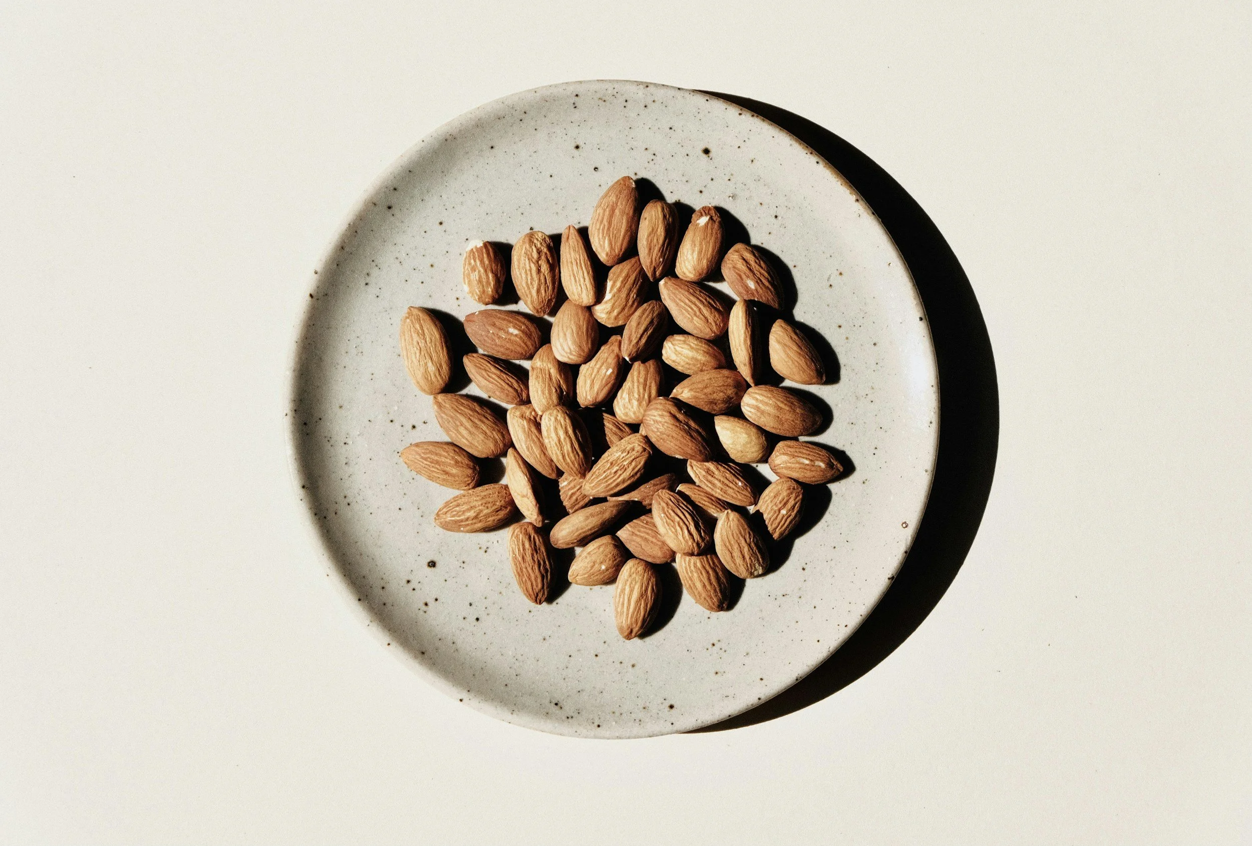 A white round plate filled with almonds placed on a white surface.