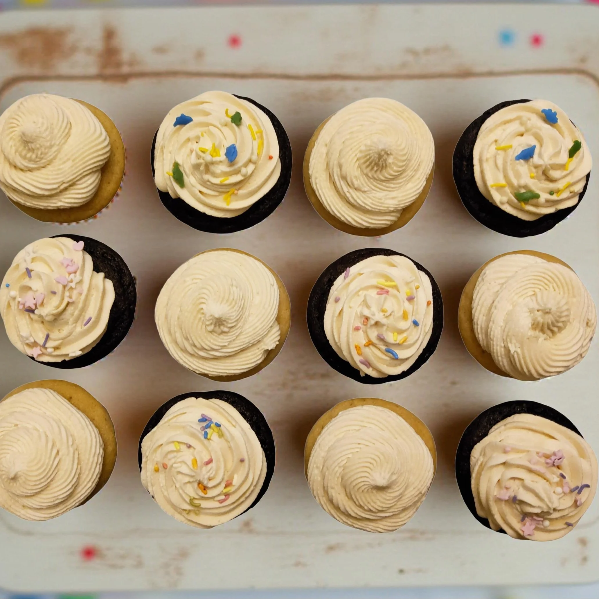 Top-down view of twelve vegan vanilla and chocolate cupcakes with white frosting and colorful sprinkles on a white wooden surface.