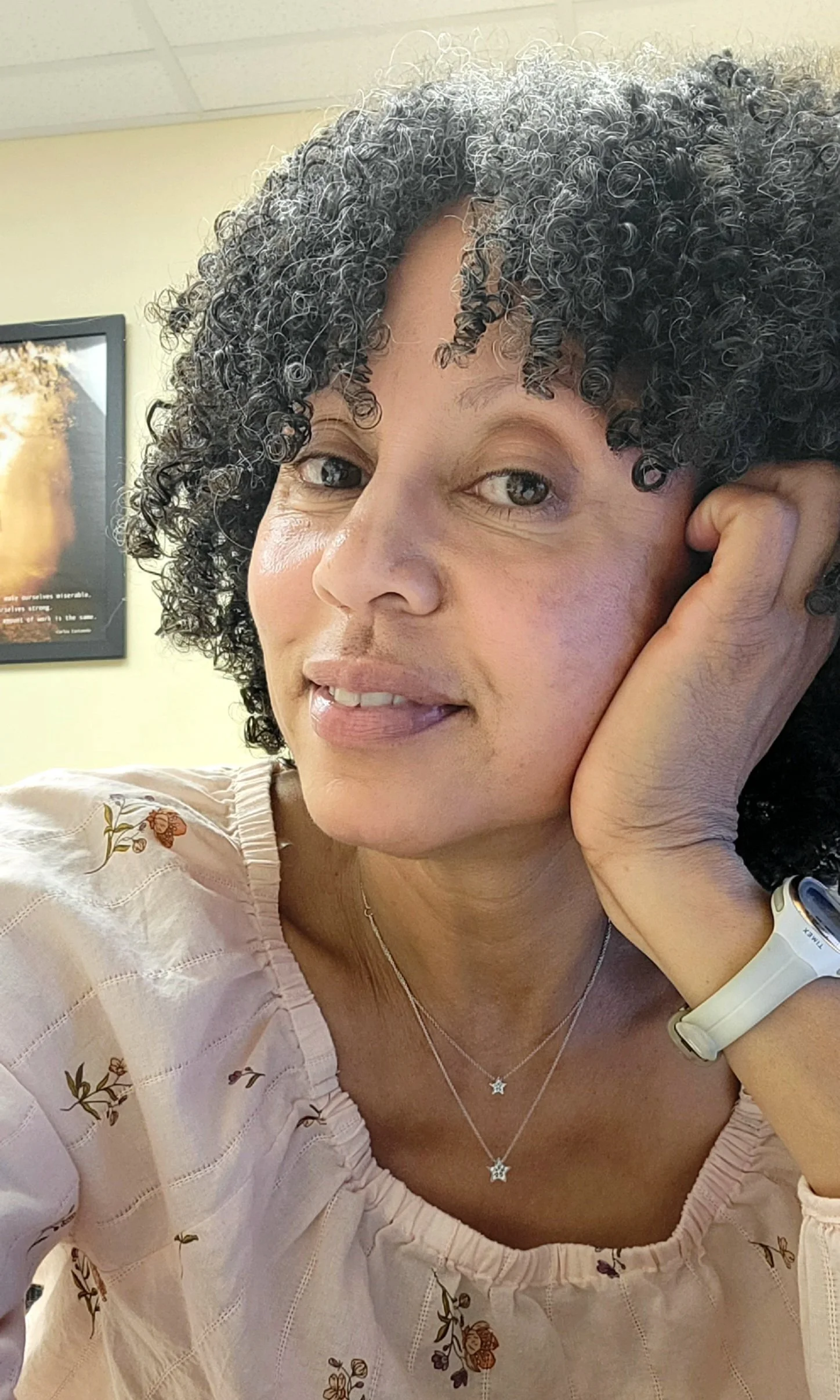 The founder and owner of Sweet Peaz Bake-House with curly hair smiling, resting her face on her hand, wearing light makeup, a light pink blouse with floral embroidery, necklaces, and a white watch, in an indoor setting