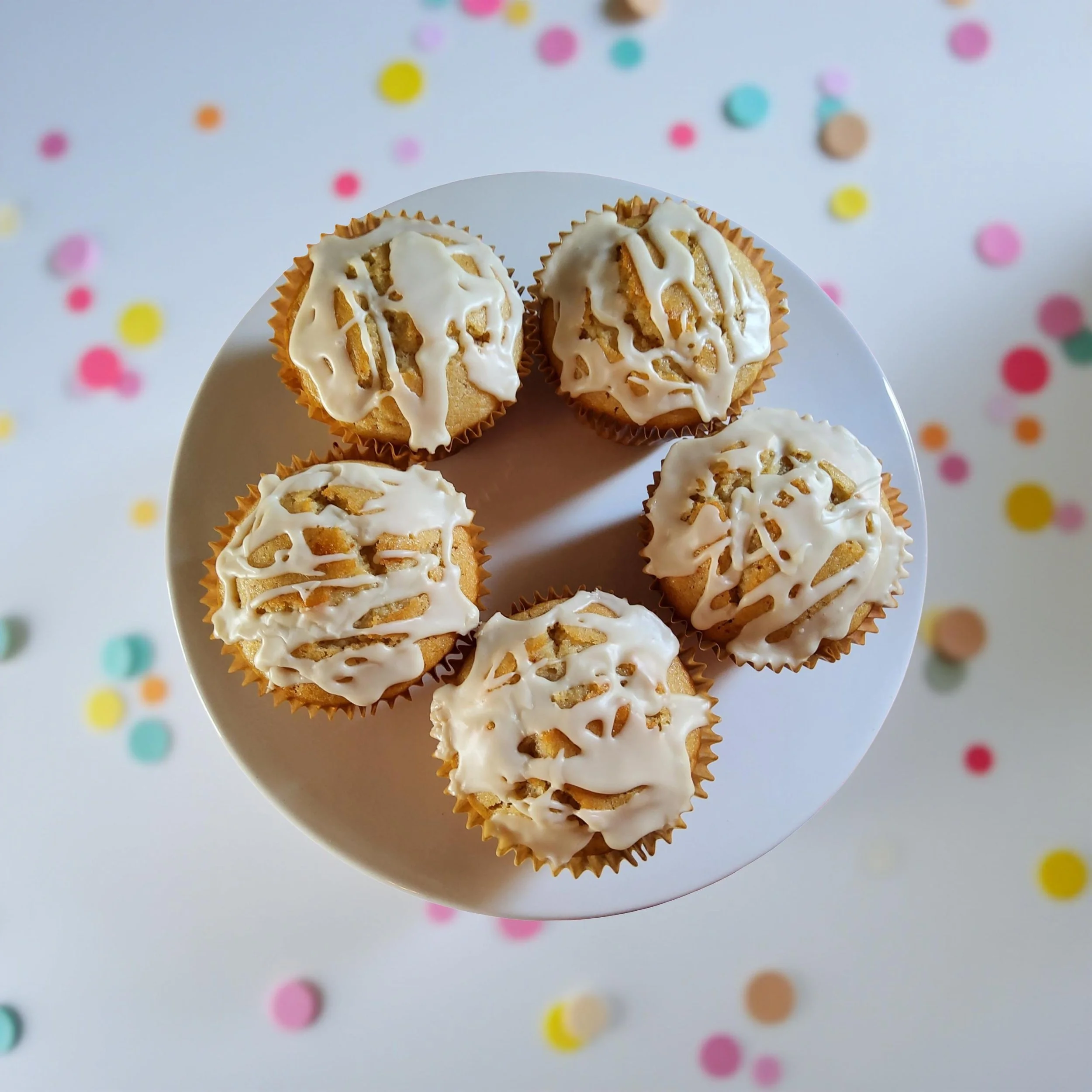 Top-down view of six jumbo vegan muffins with white icing on a white cake stand, surrounded by colorful confetti dots on a white surface.