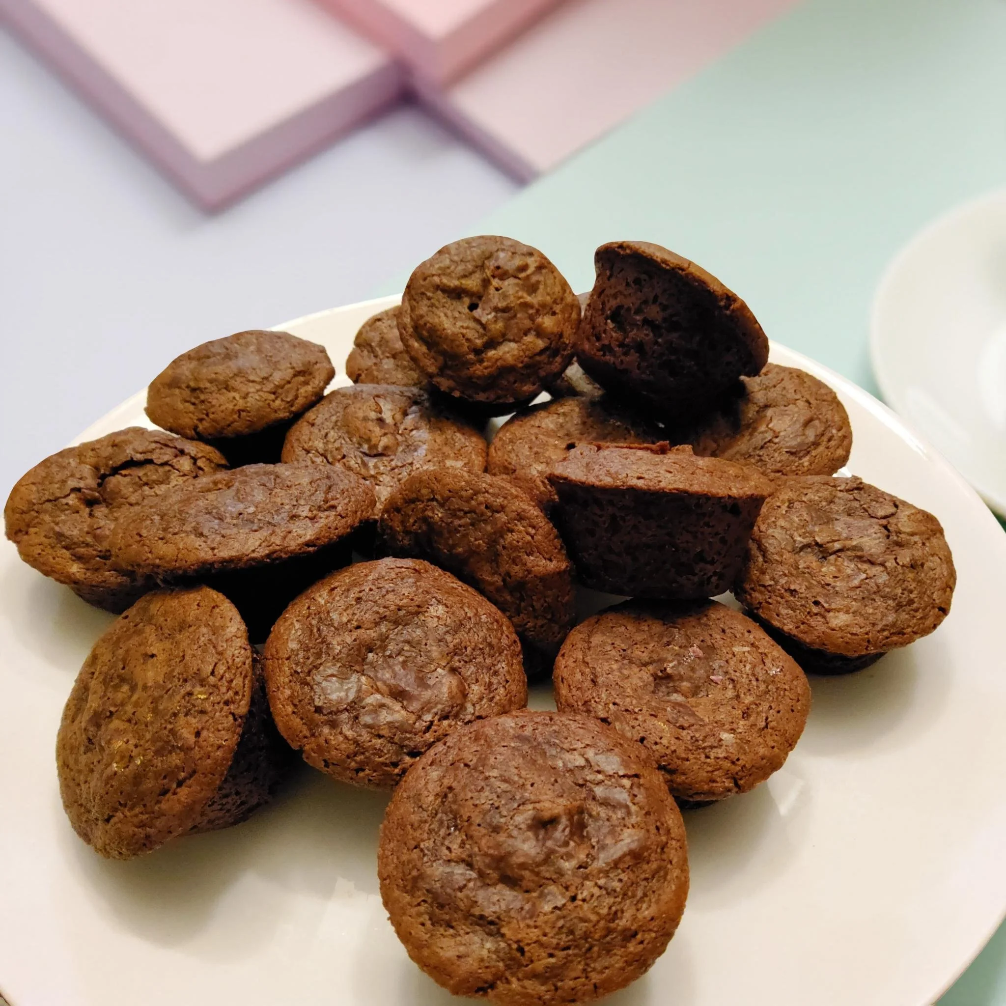 A white plate filled with vegan brownie bites, some of which are broken in half, with a box and a cup visible in the background.