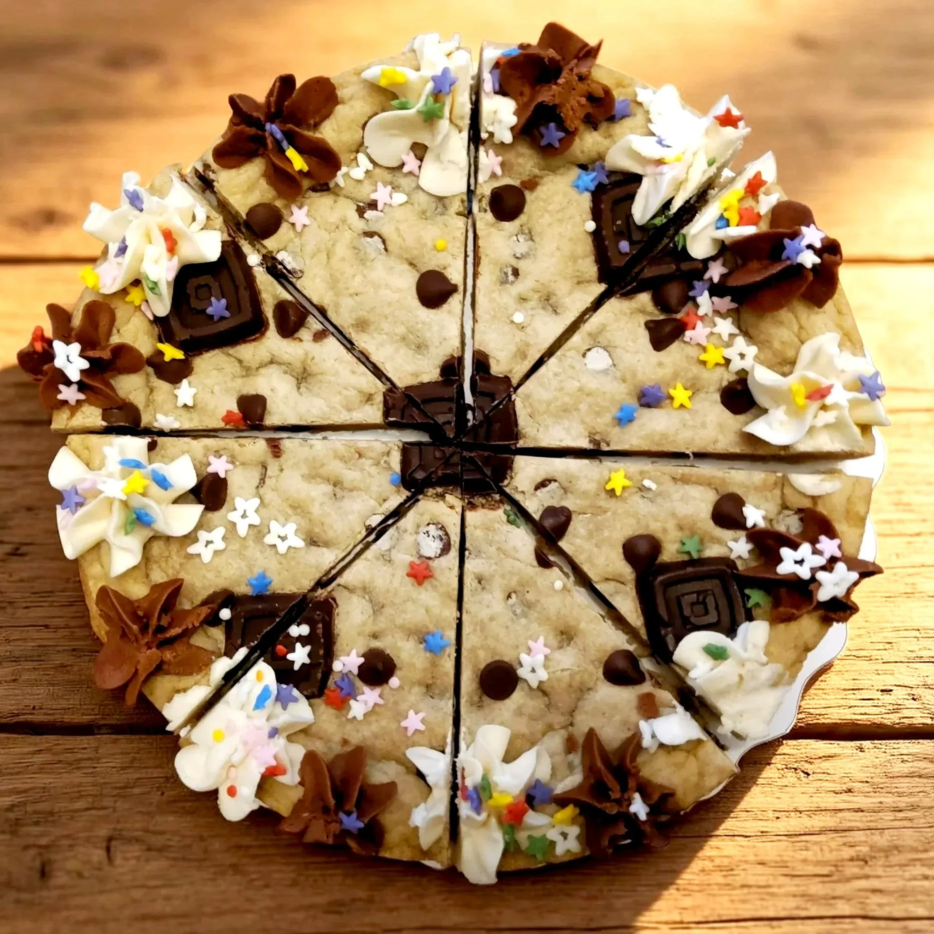 A large vegan cookie cake decorated with white and brown frosting, colorful star-shaped sprinkles, and chocolate pieces, sliced into eight pieces, on a wooden surface.
