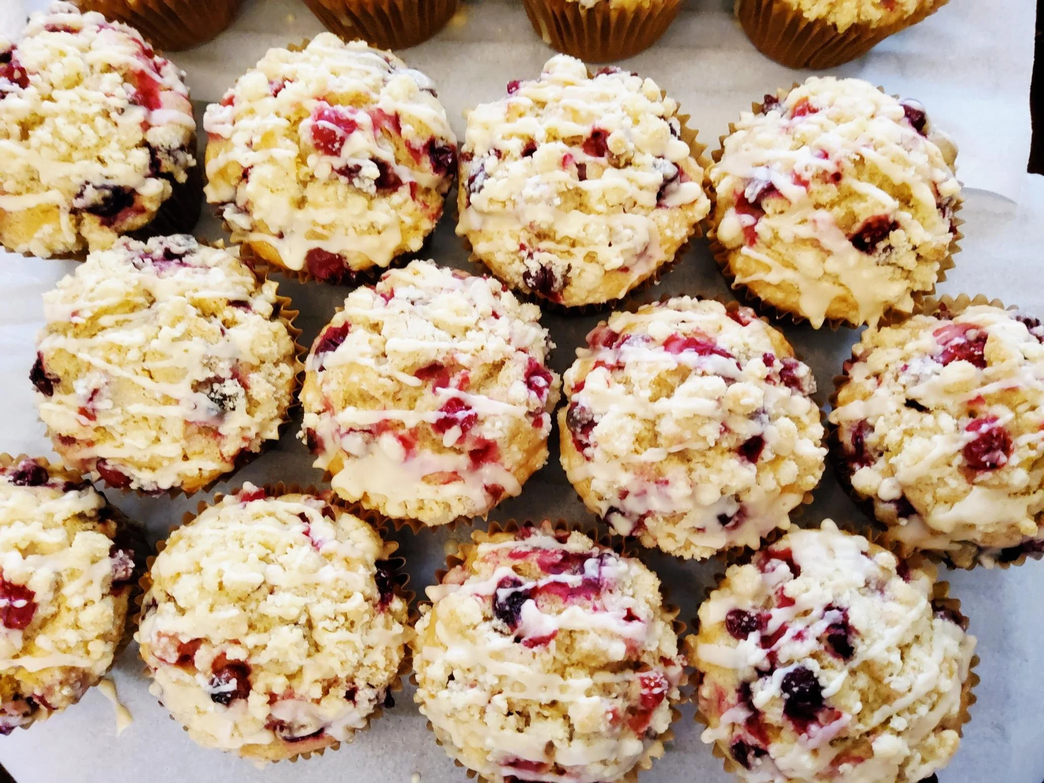 A tray of crumbly, round vegan muffins covered with white drizzle and bits of red and dark berries, placed in individual paper cups.