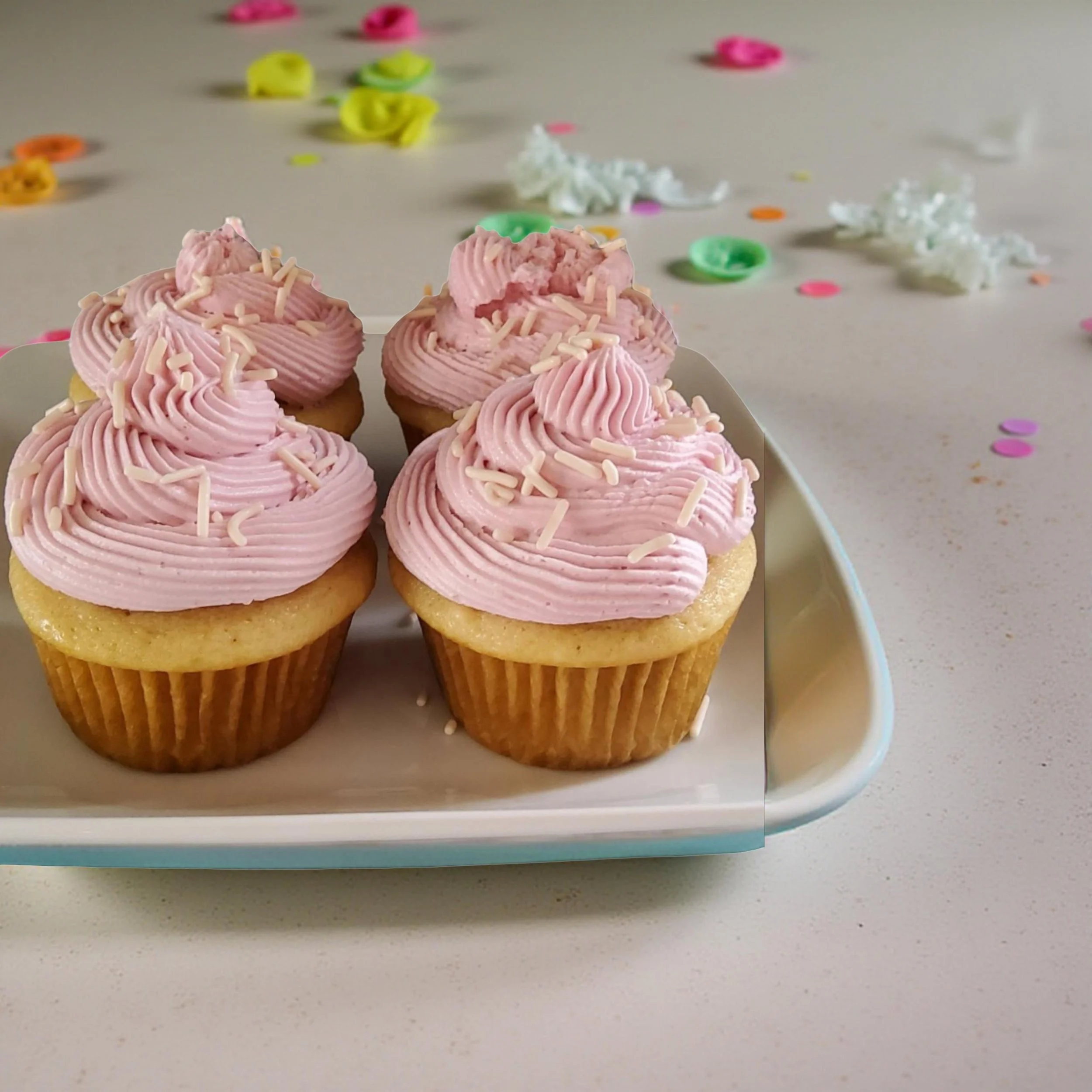 Four vegan vanilla cupcakes with pink frosting topped with white sprinkles on a white tray, surrounded by colorful confetti.