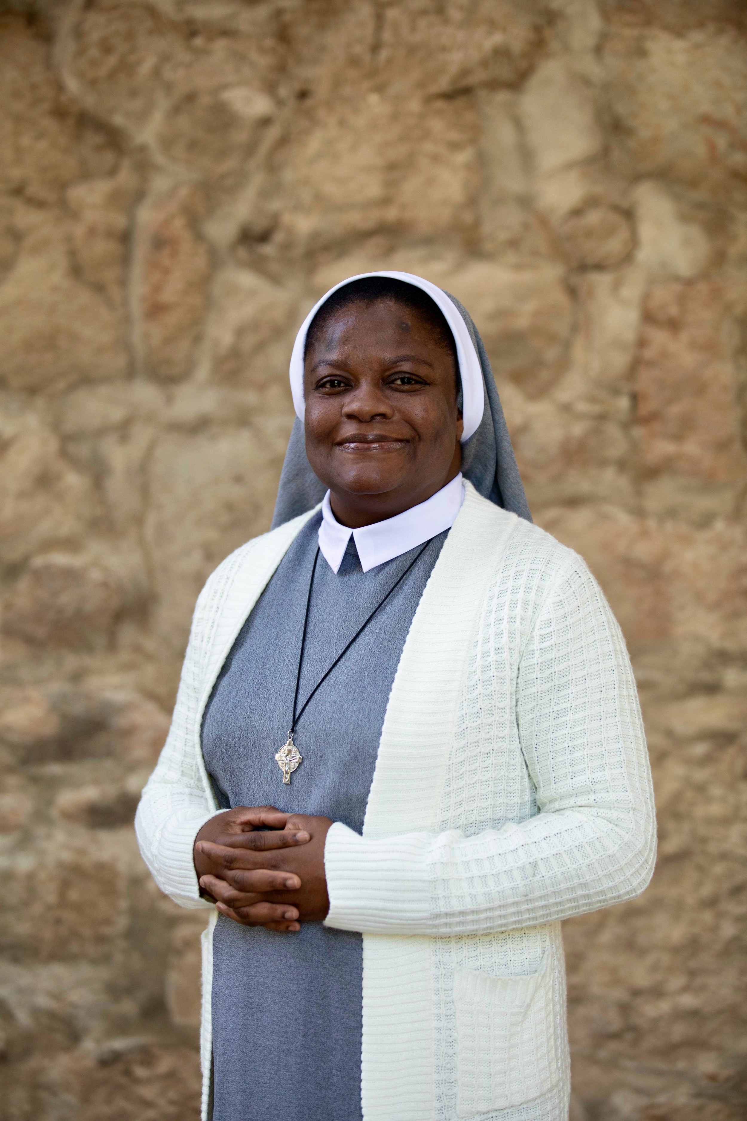 Une religieuse souriante porte une robe blanche et un voile blanc, avec un collier à lacer, devant des arbres et un ciel bleu.