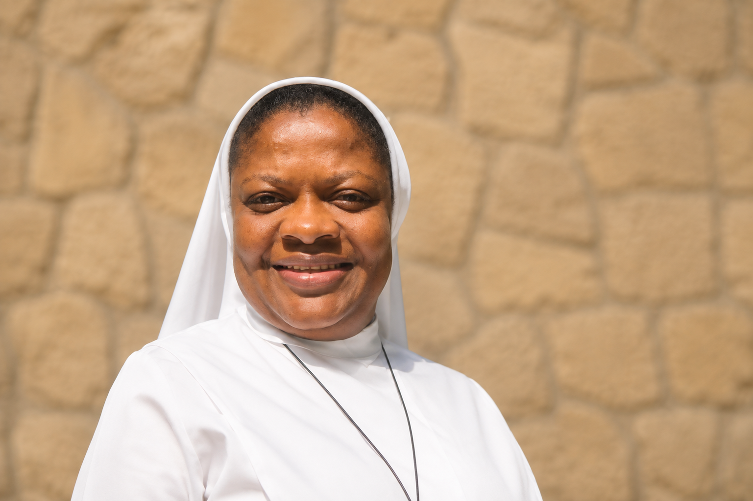 Une religieuse souriante porte une robe blanche et un voile blanc, avec un collier à lacer, devant des arbres et un ciel bleu.