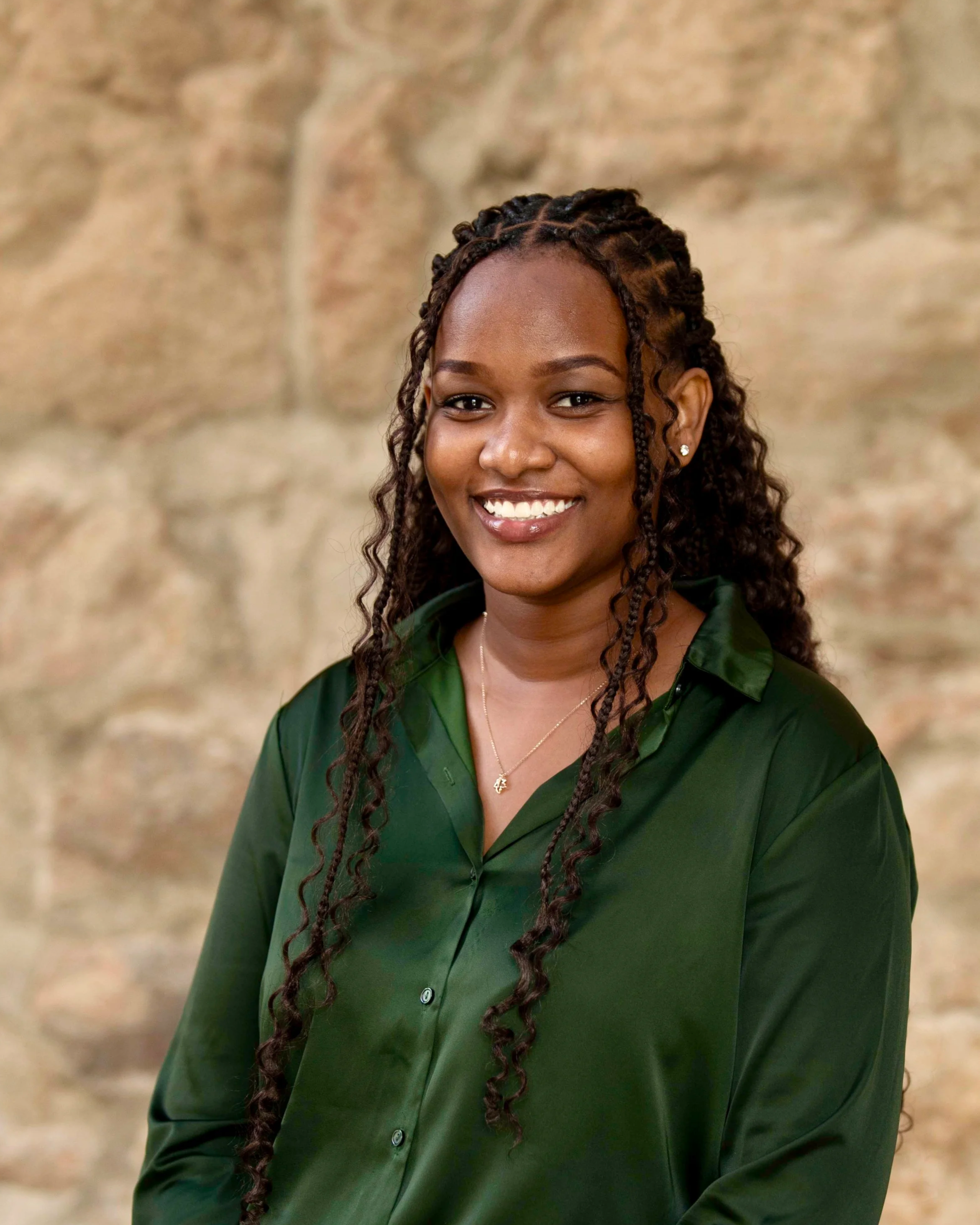 Jeune femme africaine souriante portant un pull vert, debout devant un mur en pierre.