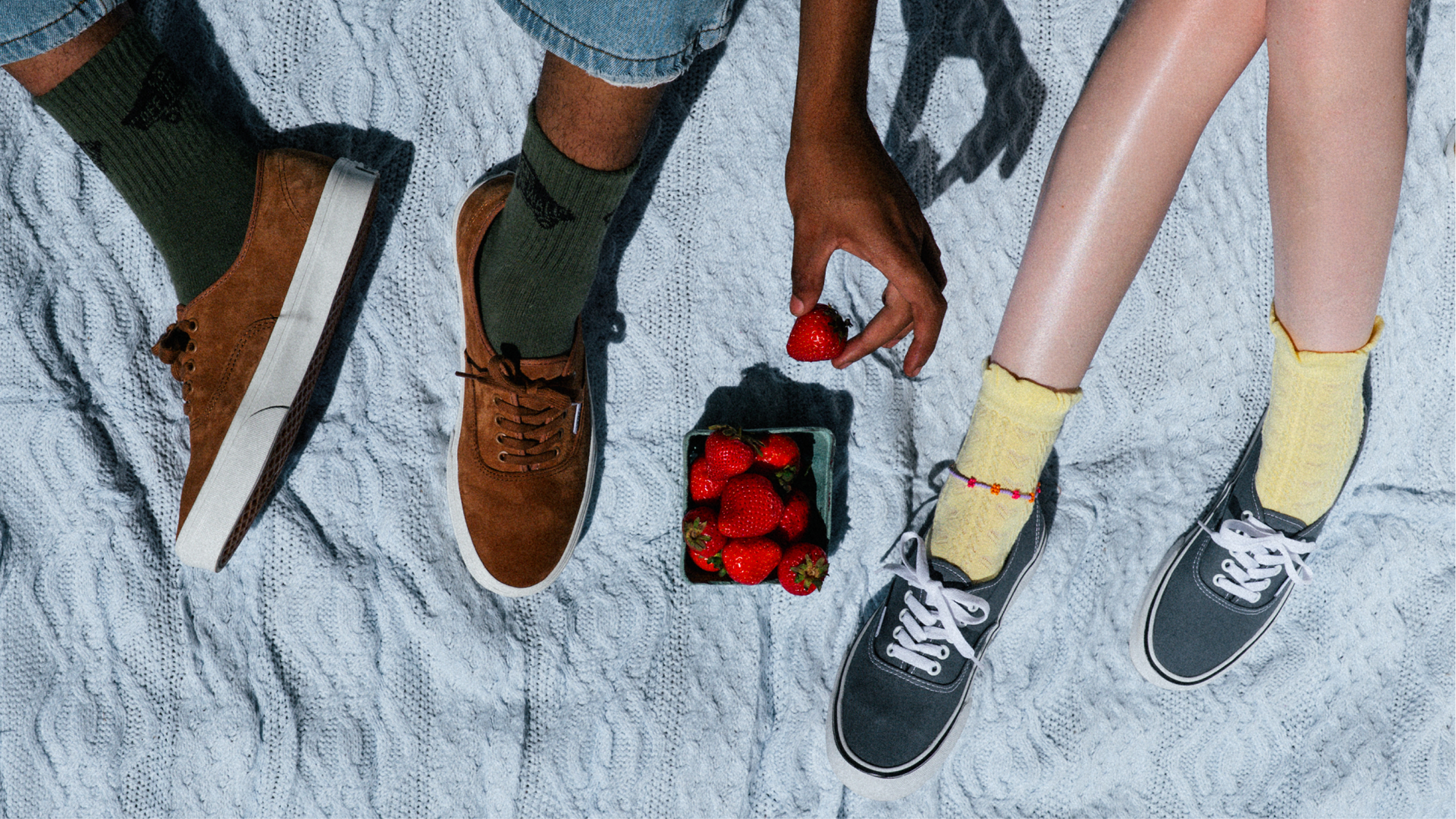 Two people sitting on a textured white blanket, one person with green socks and brown shoes, the other with yellow socks and black shoes, sharing strawberries from a container.