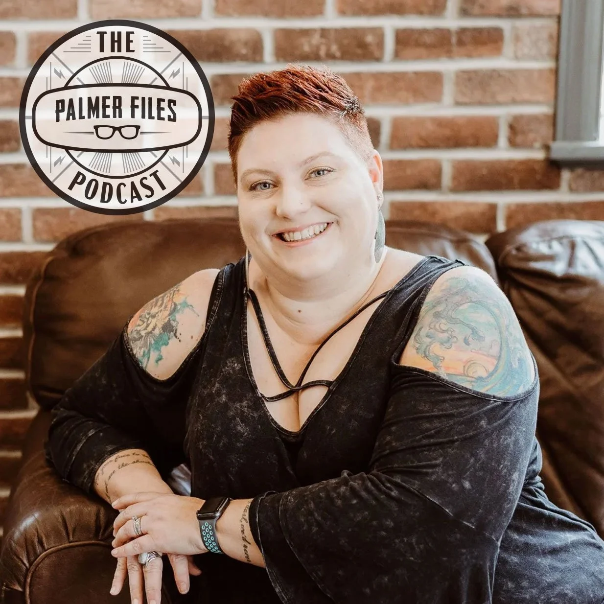 A woman with short red hair smiling while sitting on a brown leather armchair in front of a brick wall. She has tattoos on both arms and is wearing a black off-shoulder shirt, a watch, and rings, with a podcast logo in the top left corner reading 'The Palmer Files Podcast'.