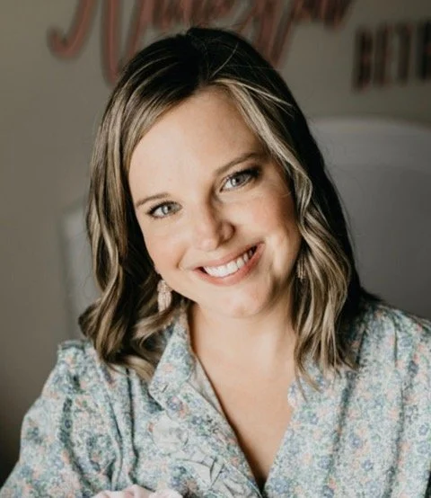 A smiling woman with wavy, shoulder-length hair, wearing large hoop earrings and a floral blouse, stands outdoors in front of a blurred background of trees.