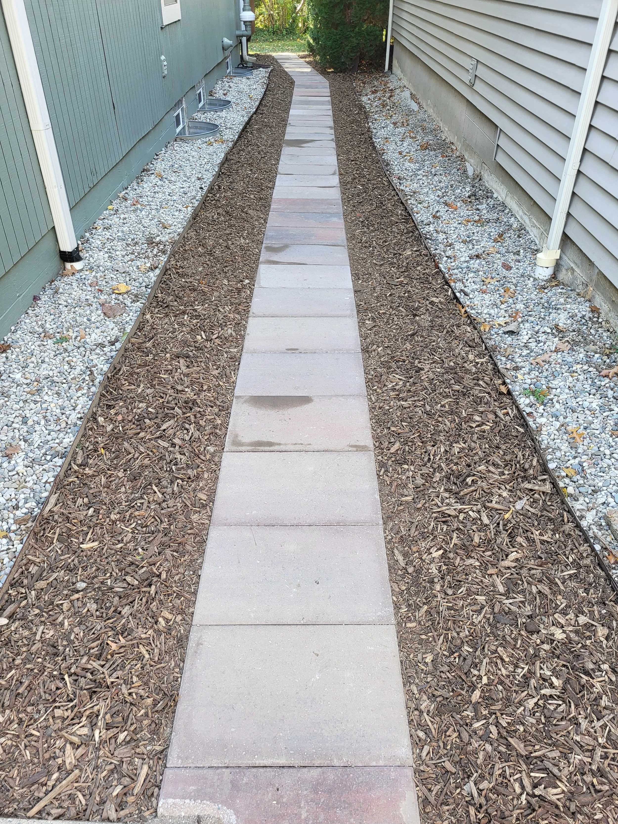 A narrow concrete walkway with steps in between, bordered by mulch and gravel on both sides, running between two houses.