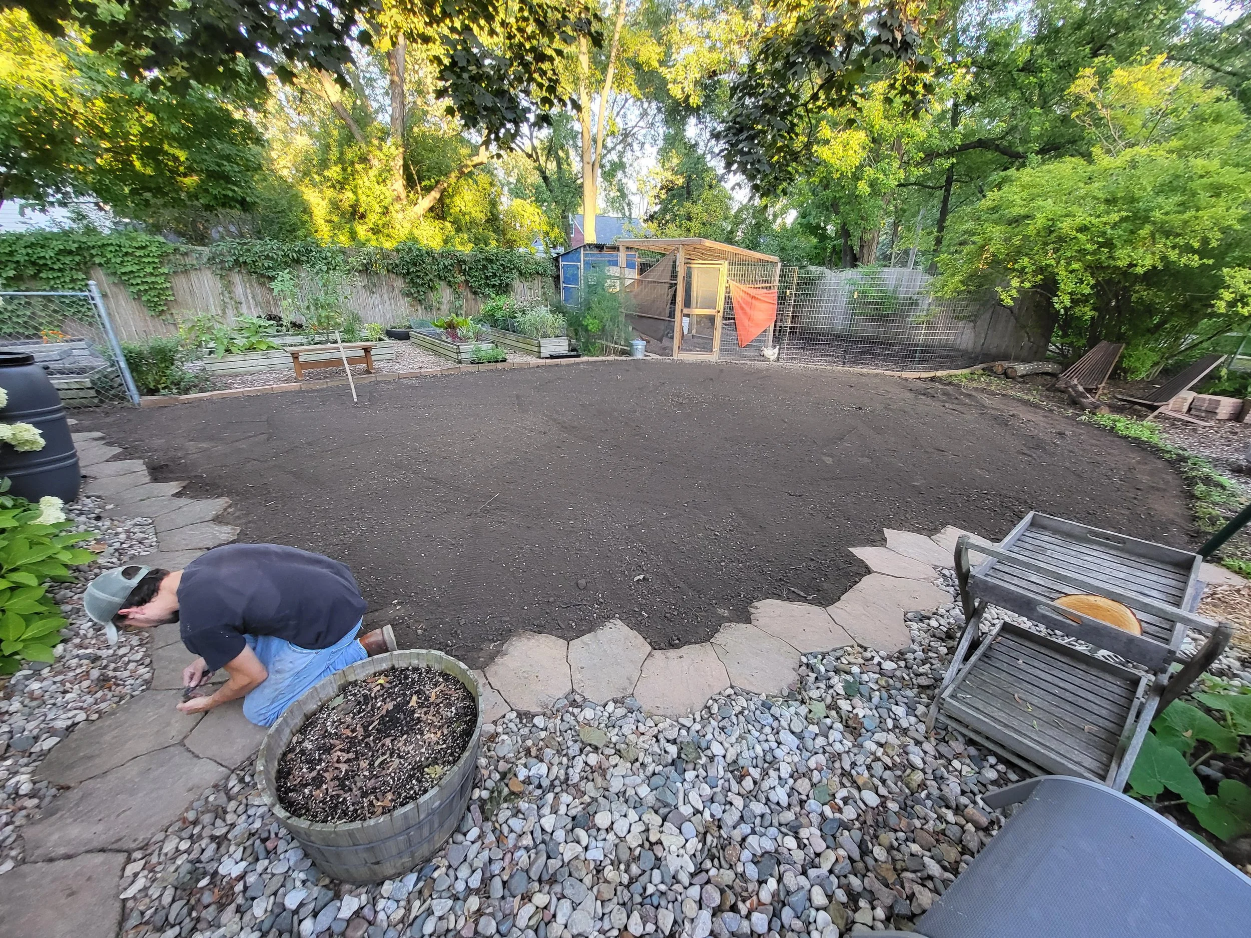 A person kneeling on a stone pathway in a backyard, planting or tending to soil in a corner. The yard has a large, cleared section of dark soil, surrounded by gravel and stones, with trees and a small wooden shed in the background.