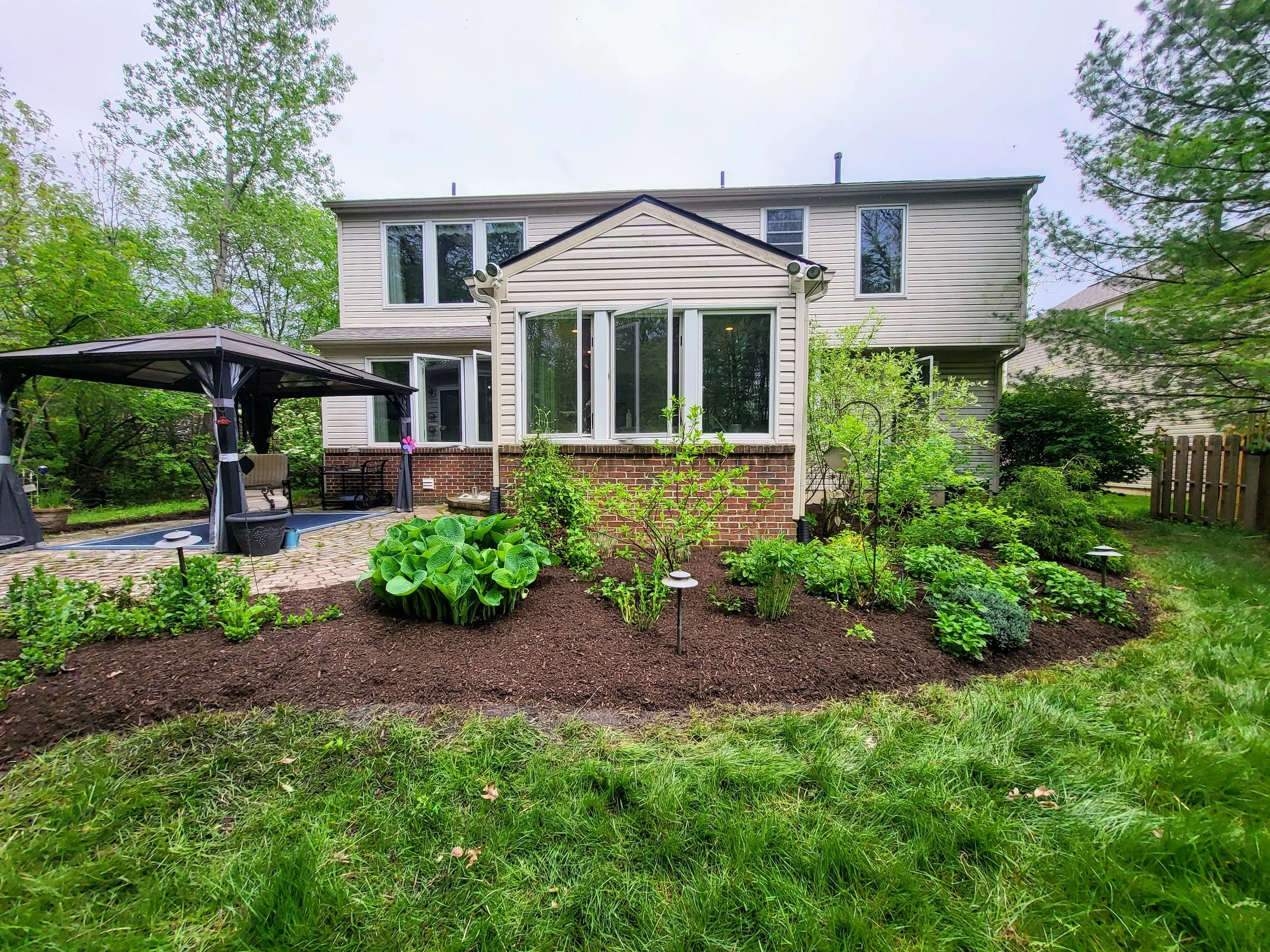 Backyard garden neatly trimmed with a variety of green plants, a brick patio, a black gazebo, and a two-story house with white siding and large windows.