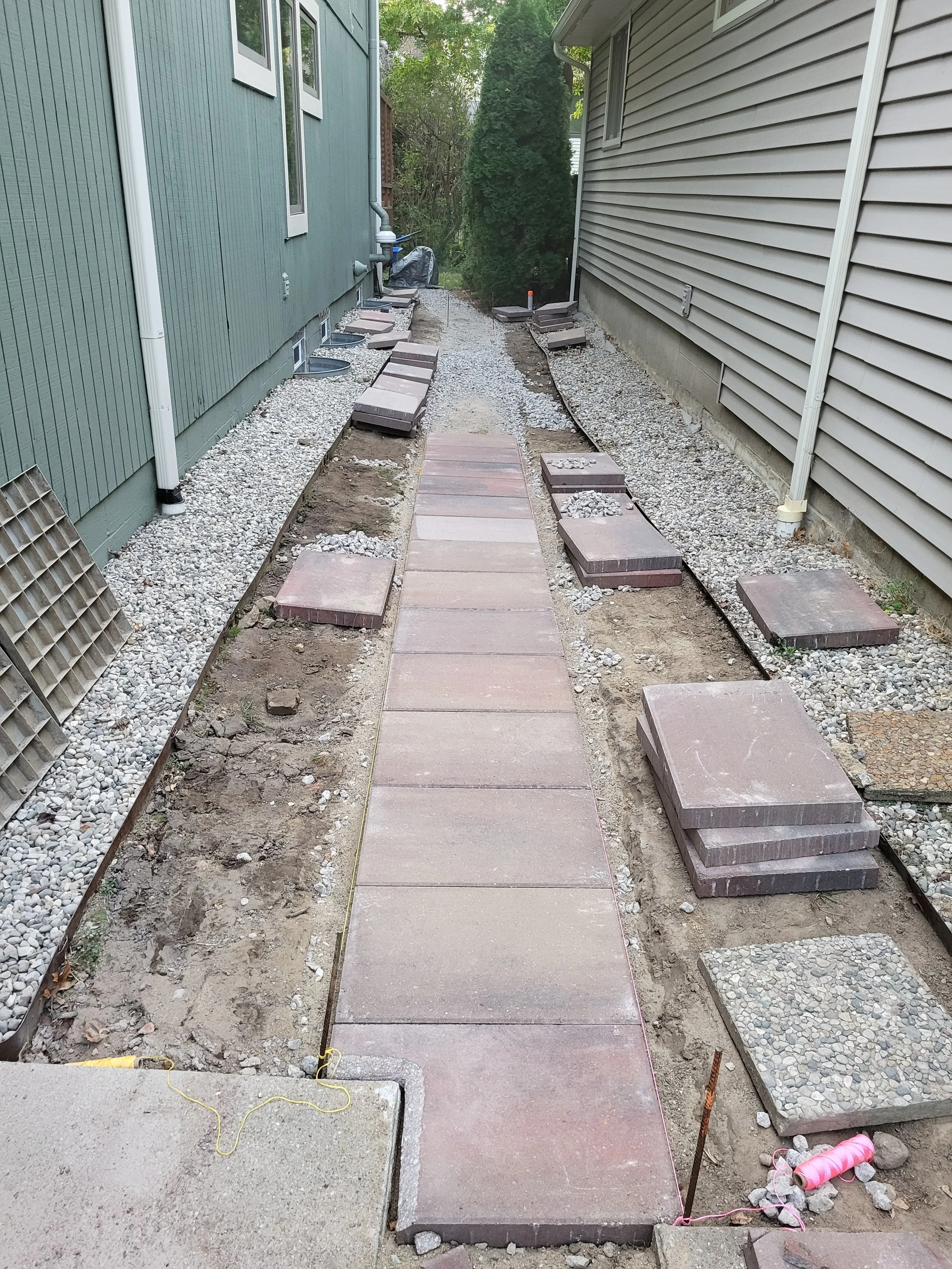 A narrow walkway under construction between two houses, with new concrete or stone pavers being installed, and construction materials and equipment nearby.