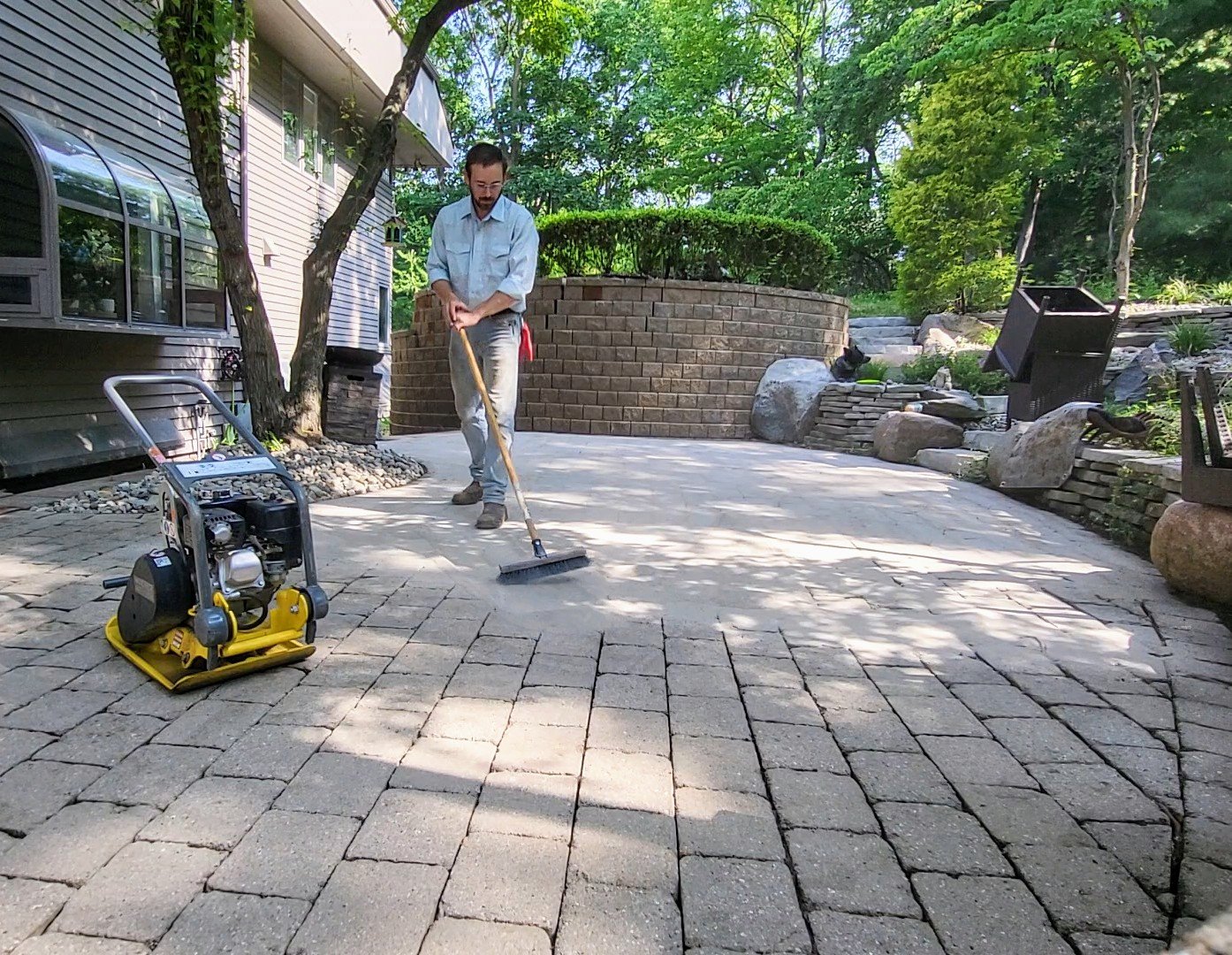 A man using a tool to work on a brick patio outdoors, with a machine nearby on the ground and lush green trees and stone landscaping in the background.