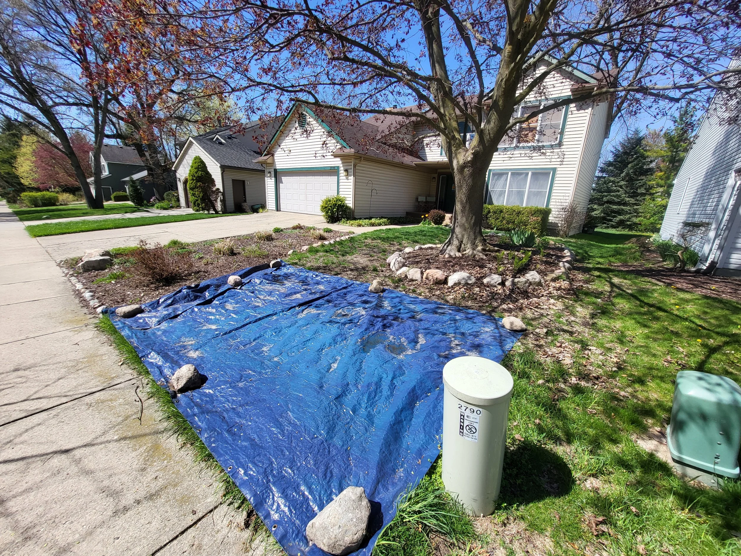 A landscaped front yard with a large tree surrounded by a stone border. A blue tarp covered with rocks is spread on the grass near the sidewalk. A white utility box with the number 2790 is on the grass next to the tarp. The house in the background has white siding, a two-car garage, and a small porch.