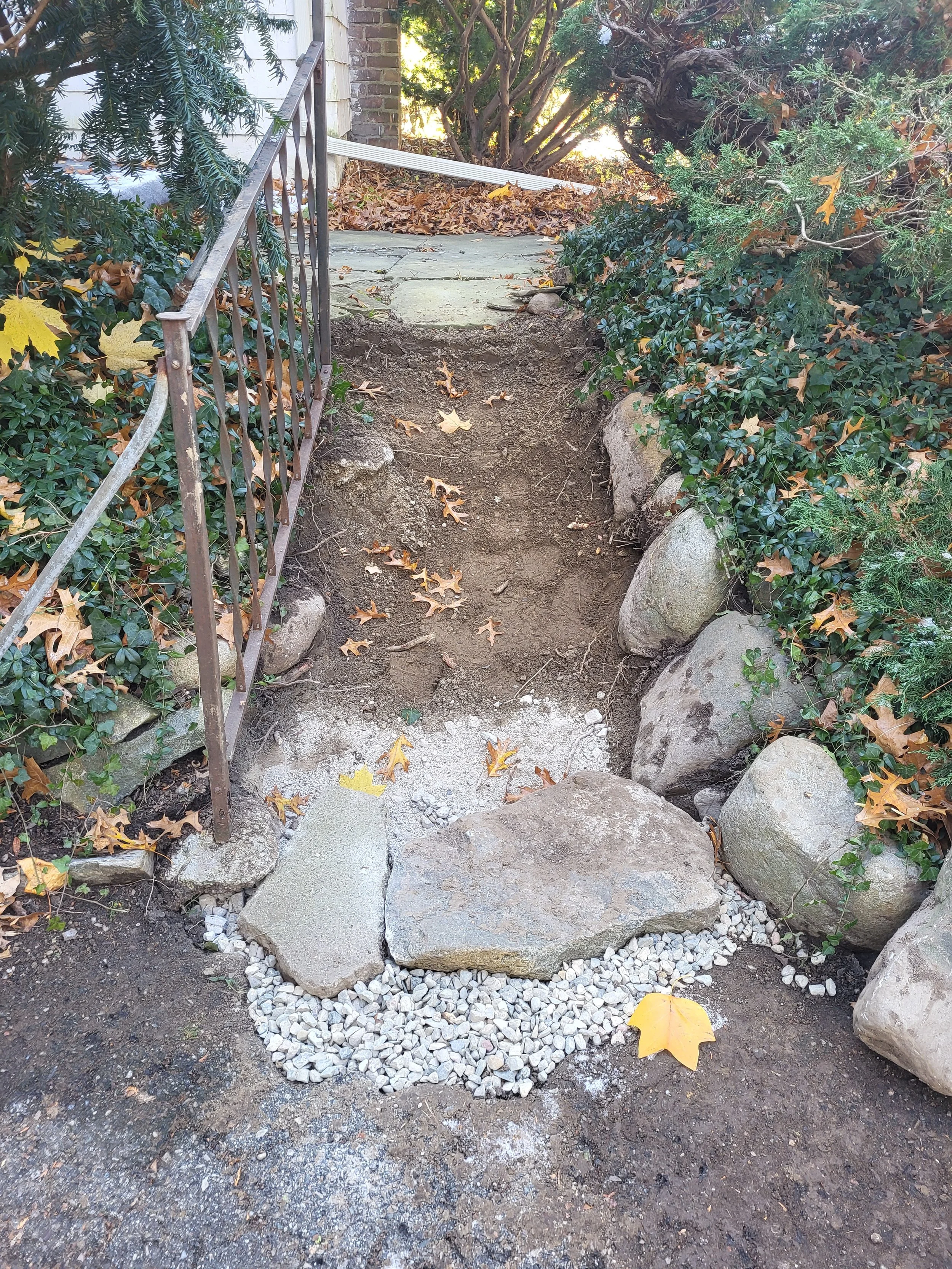A garden pathway under construction with exposed dirt, bordered by large rocks and small white gravel, next to a metal railing and surrounded by greenery and fallen autumn leaves.