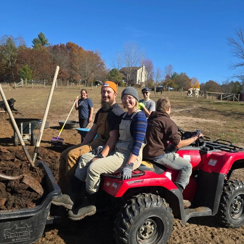 People working on a farm or garden, sitting on a red utility vehicle, with others in the background raking and digging soil, and a house with trees and a blue sky in the background.