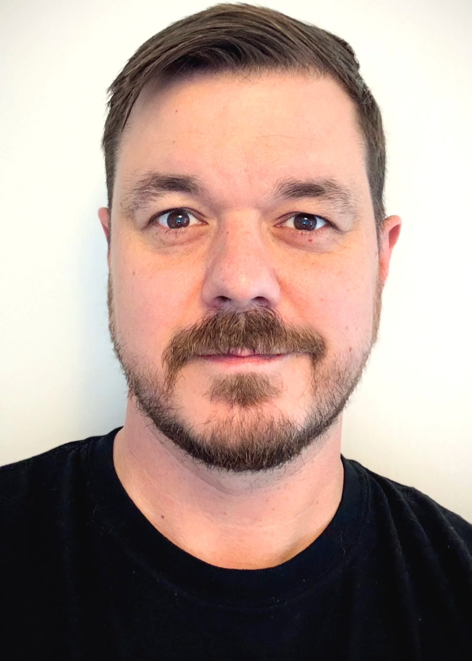 Close-up of a smiling man with short brown hair, facial hair, wearing a black shirt, dark eyes, and light skin, standing against a plain white background.