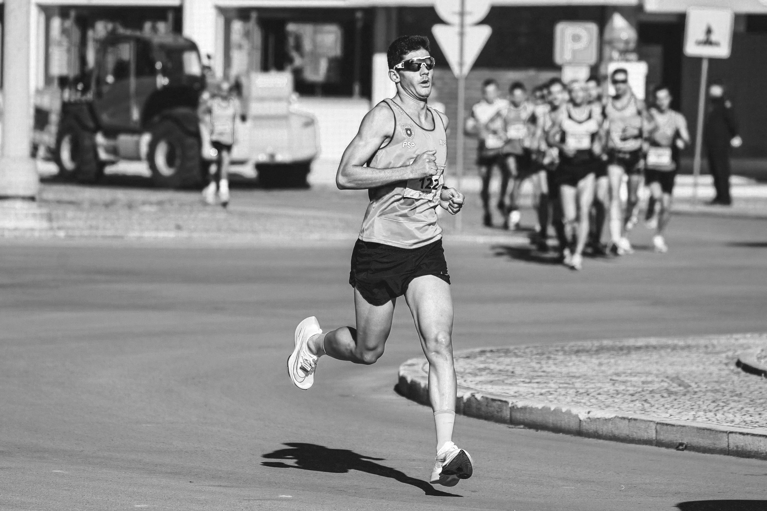 A male runner wearing sunglasses, a tank top, and running shorts is participating in a race on a city street. There are other runners in the background, along with street signs and a bus.