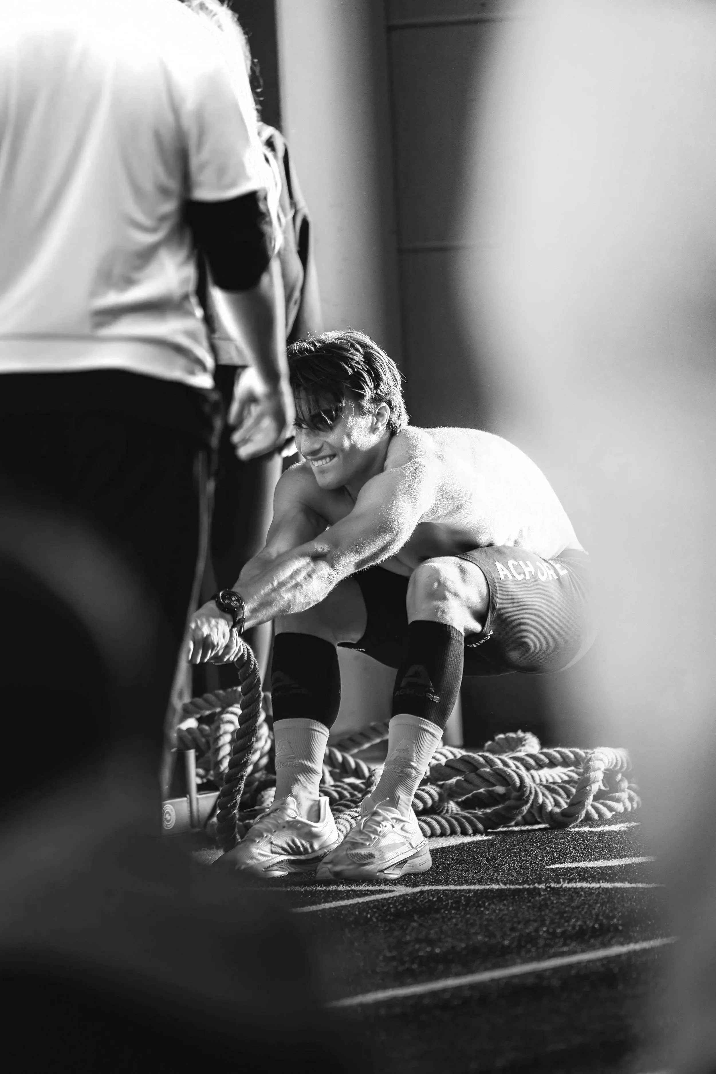 A young man is smiling and adjusting his sports shoes while crouching on the floor, with a coach or trainer standing nearby. They are in what appears to be a training or gym environment. The image is in black and white.