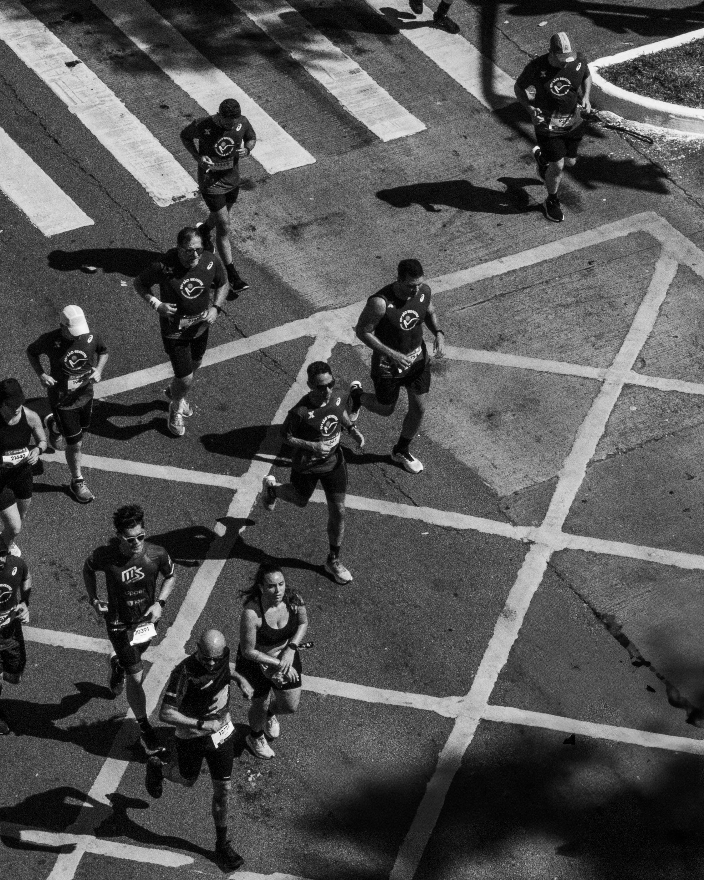 Black and white photo of marathon runners crossing an intersection on a city street at night, shadows cast on the pavement.