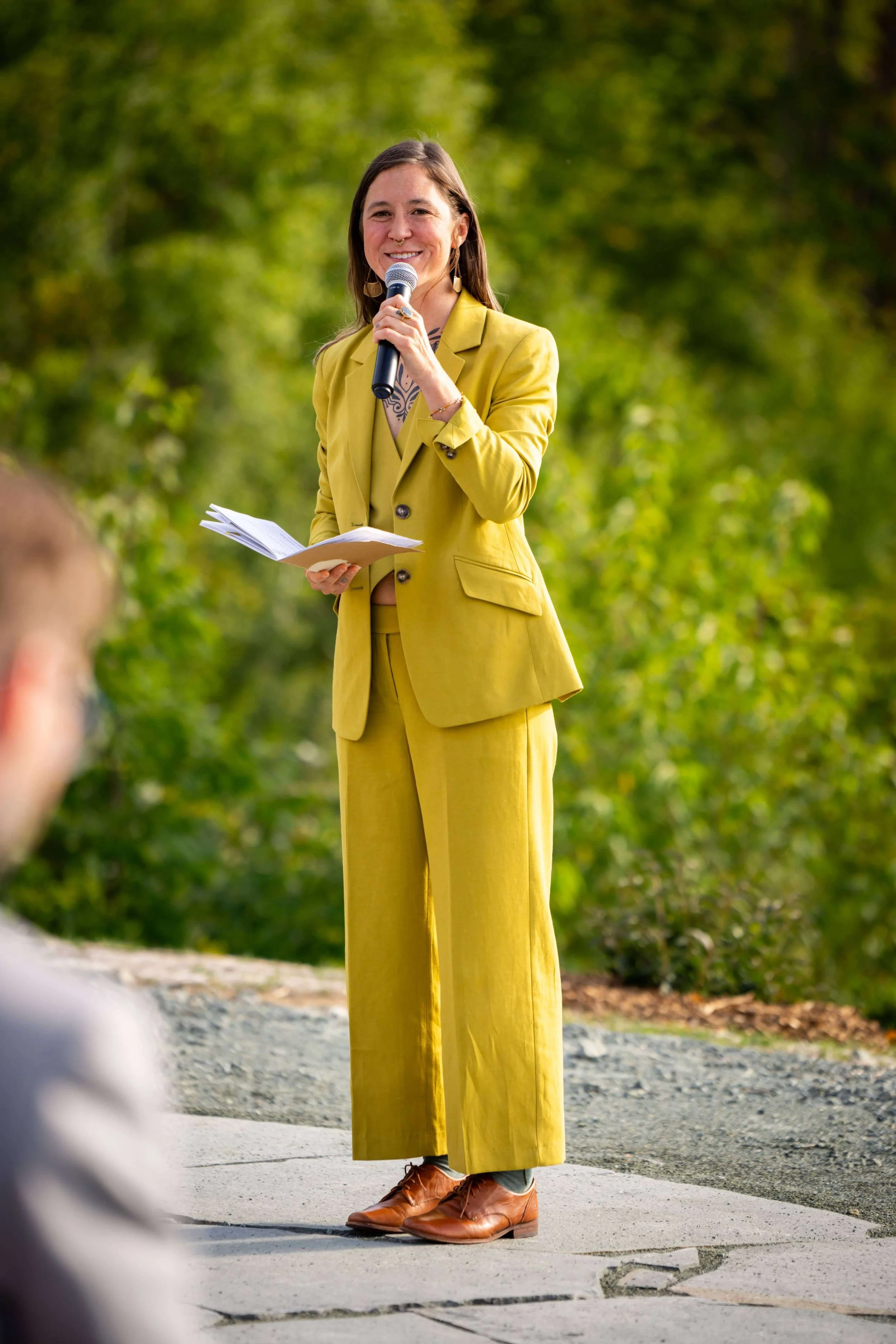 speaker standing on a stone patio in a lime green suit speaking into a microphone, smiling, and holding a program