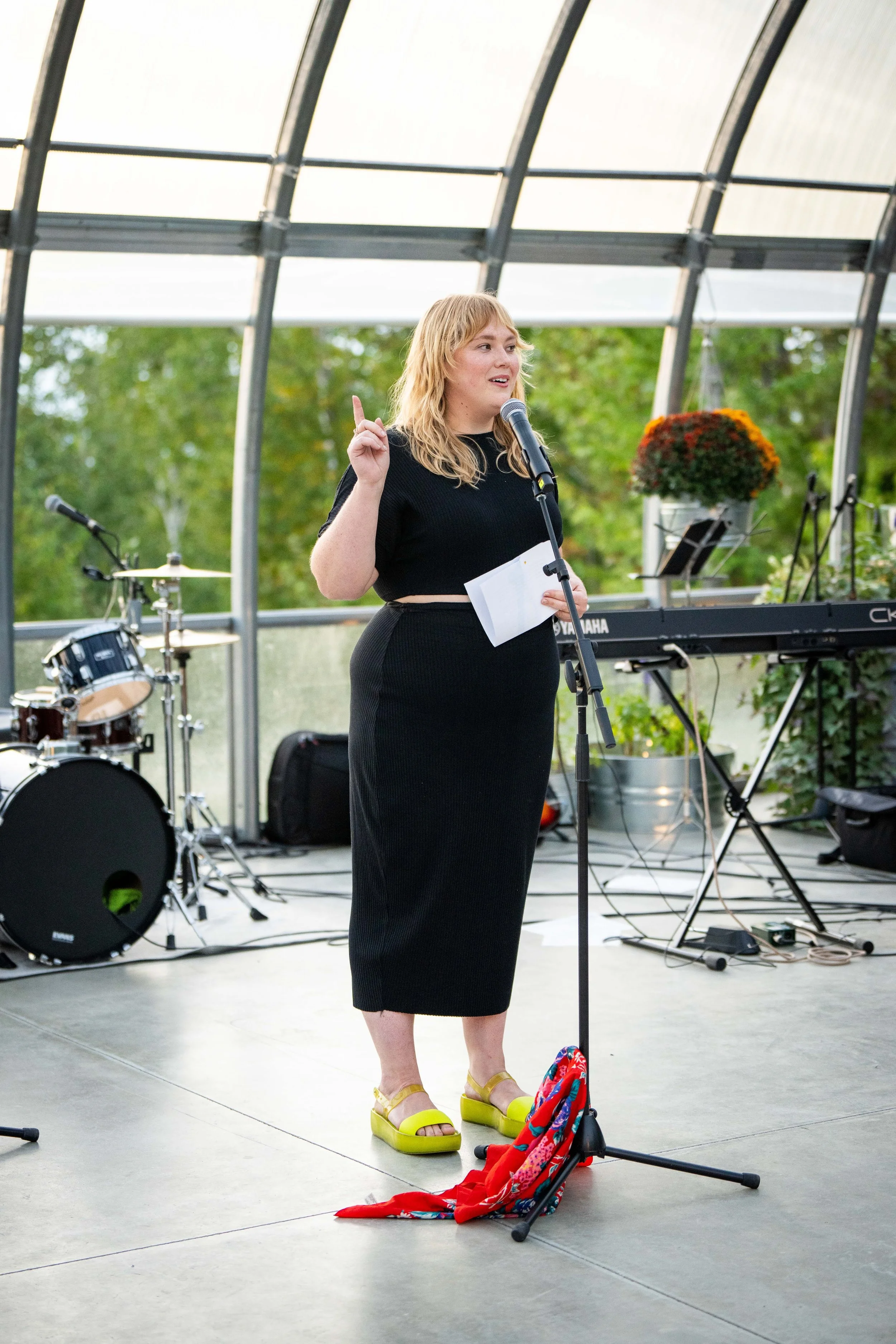 speaker standing in a greenhouse with a microphone, wearing a black skirt and top and pointing in the air