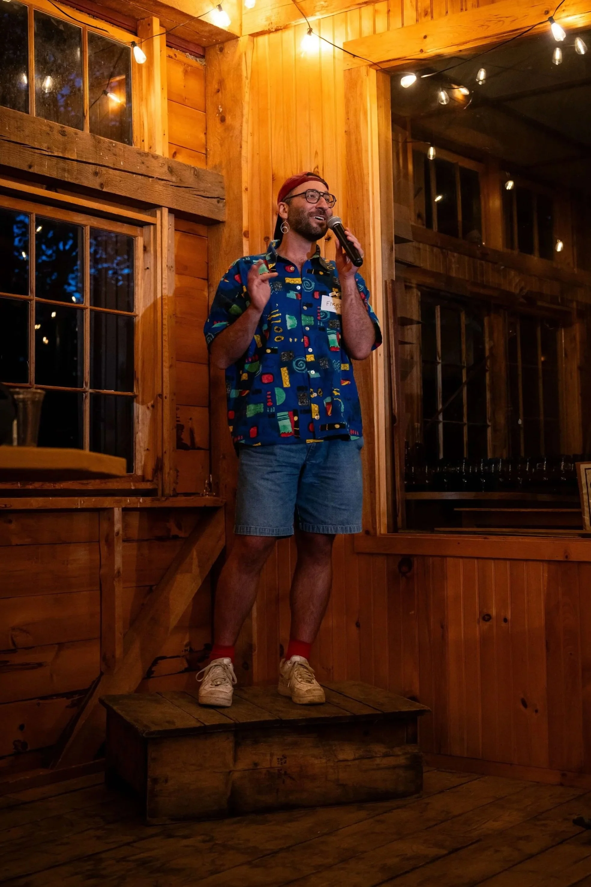 speaker with blue shirt in a wooden building standing on a box speaking into a microphone and smiling
