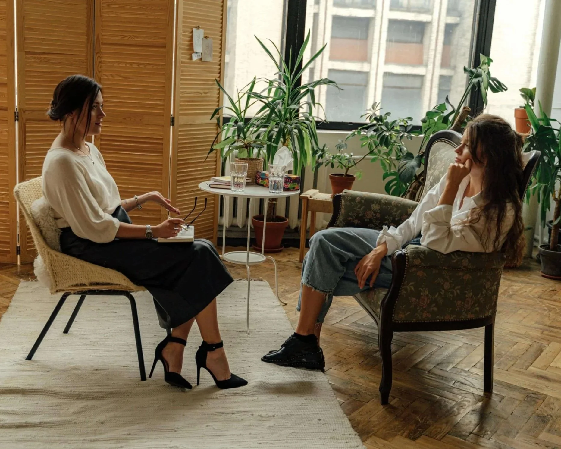 Two women having a conversation in a cozy indoor space. One woman, with dark hair in an updo, is seated on a woven chair, wearing a white blouse and black skirt with high heels. The other woman, with long wavy hair, is seated on a vintage armchair, wearing a white shirt and ripped jeans. The background features large windows, indoor plants, wooden floors, and a wooden privacy screen.
