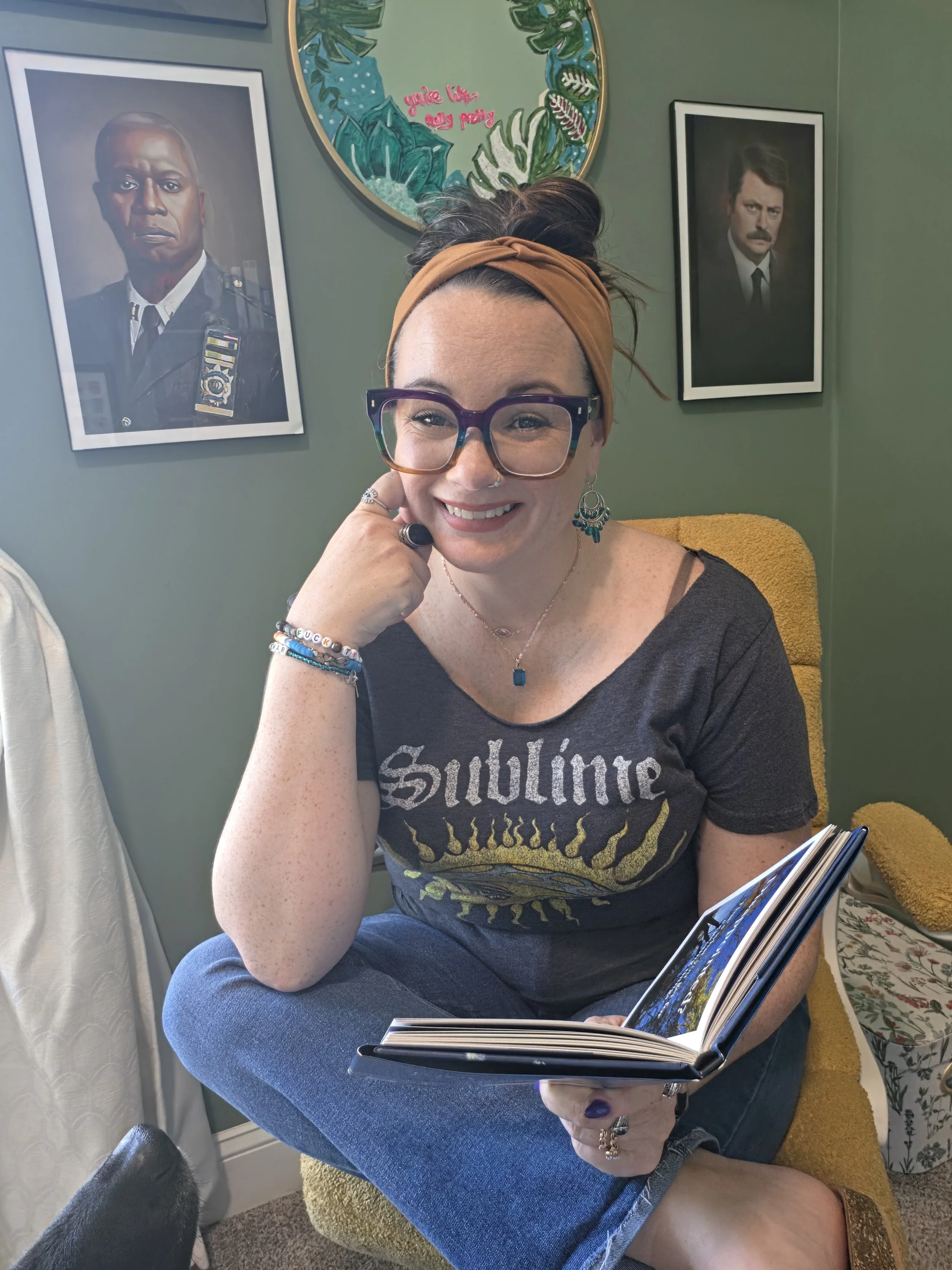 A woman with glasses, earrings, and a headband sitting on a yellow chair, smiling and holding an open book or magazine. Behind her are framed photographs on a green wall, a person in a police uniform on the left, and a man with a mustache in a suit on the right. There is also a round mirror decorated with leaves and a pink phrase that says 'you're like really pretty'.