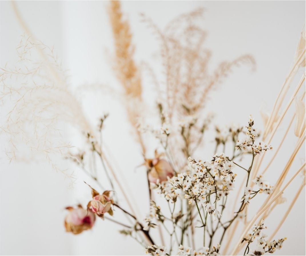 A close-up of a dried flower bouquet with small white flowers and pink buds, set against a light, neutral background.