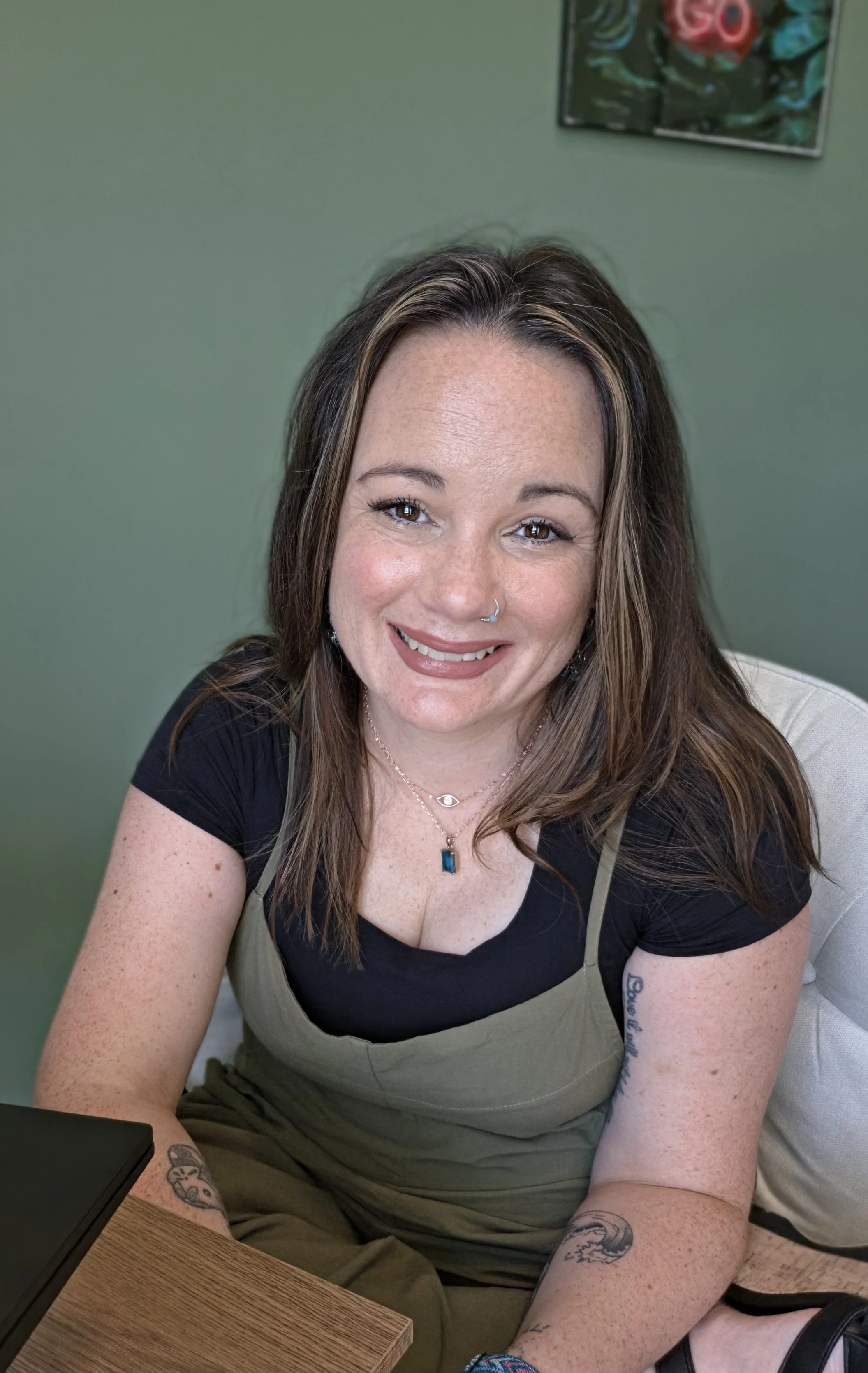 A smiling woman with long brown hair, wearing a black top and beige apron, sitting at a wooden table with tattoos on her arms. There is a green wall and a colorful poster in the background.