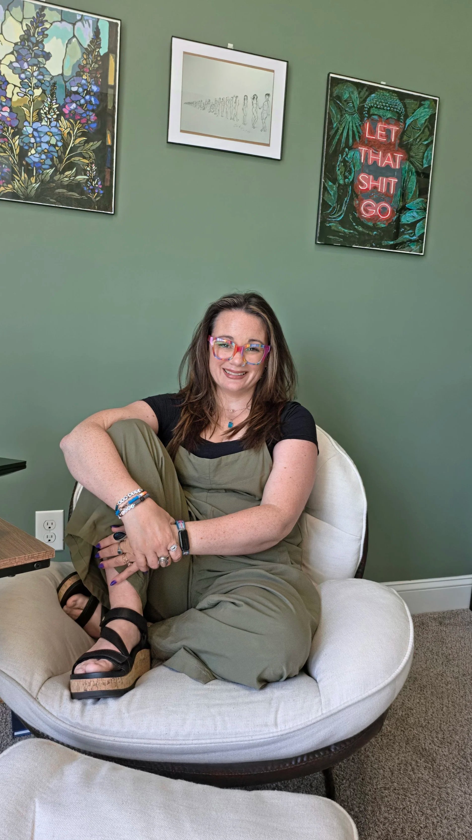 A woman sitting comfortably on a round cushioned chair in a room with green walls, wearing colorful glasses, a black shirt, and loose greenish pants. She has long brown hair and is smiling. Behind her are framed art pieces on the wall, including a colorful floral painting, a minimalist line drawing of people, and a vibrant poster with a nature scene and the phrase 'LET THAT SHIT GO' in glowing red letters.