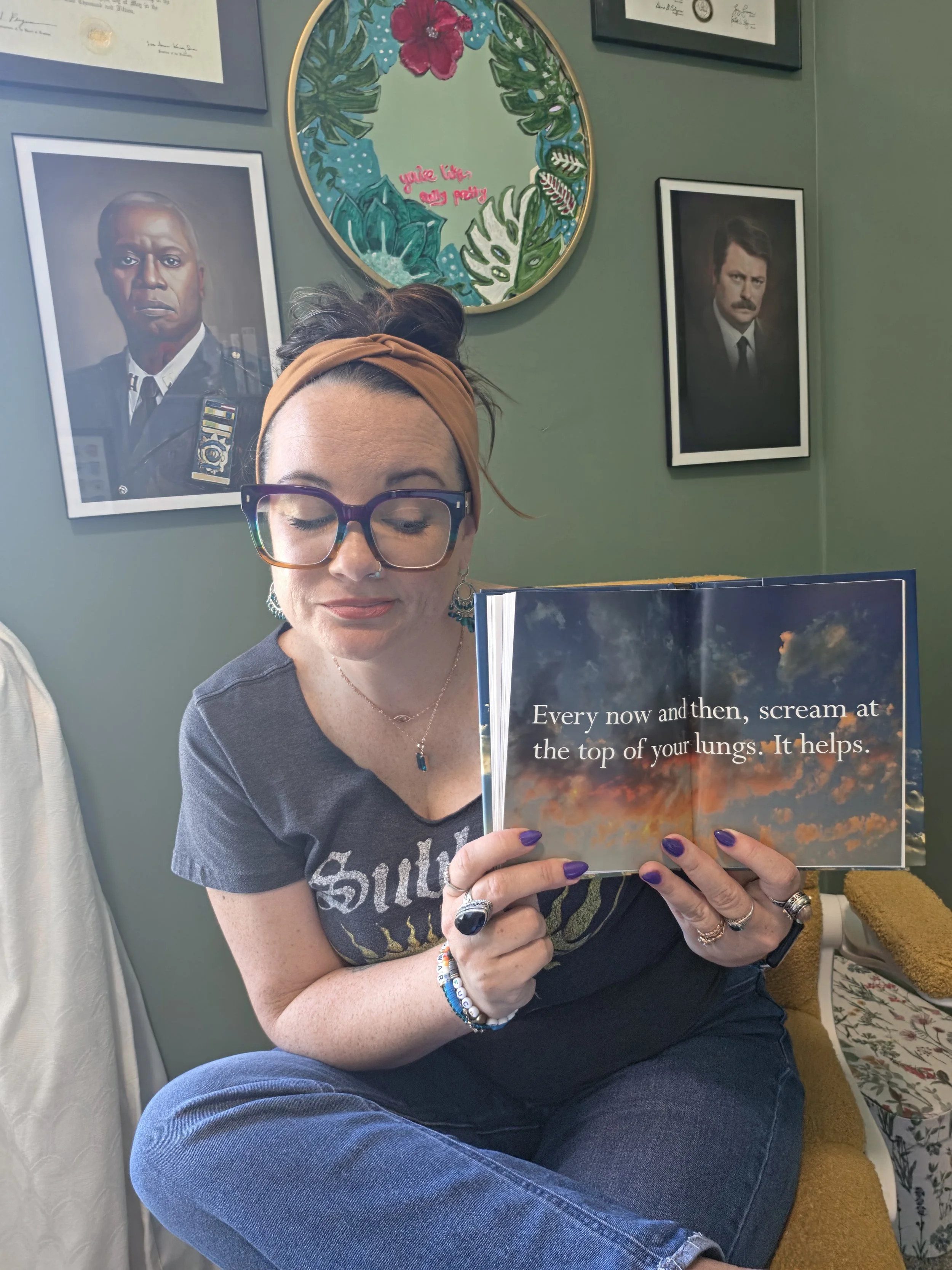 A woman with glasses and a headband sitting cross-legged on a yellow chair, holding a book with a colorful sunset sky on the cover. The book's text reads: "Every now and then, scream at the top of your lungs. It helps." She is wearing a dark graphic T-shirt and has multiple rings, bracelets, and a necklace. The background features framed portraits and artwork on the wall.