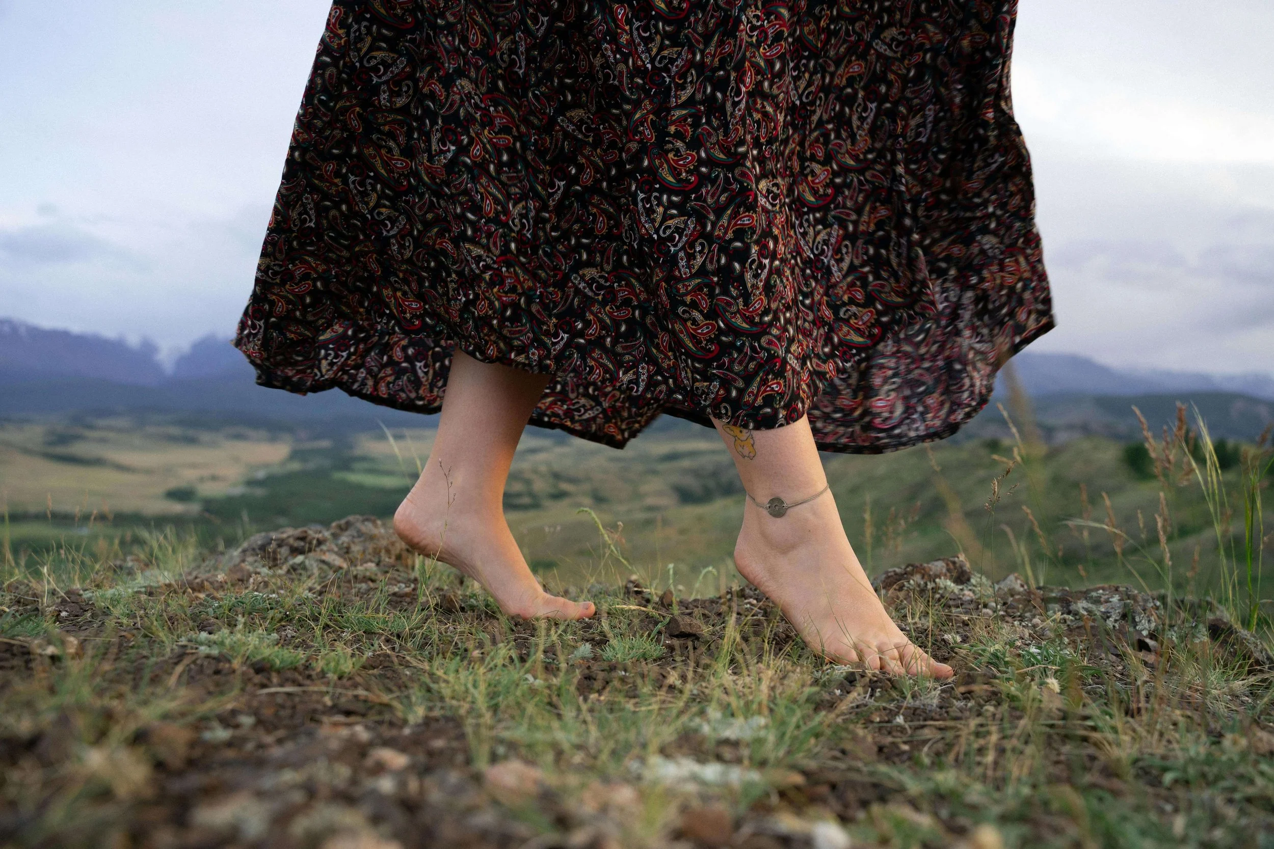 Person with a dress walking barefoot through a grassy outdoor area with mountains and cloudy sky in the background.