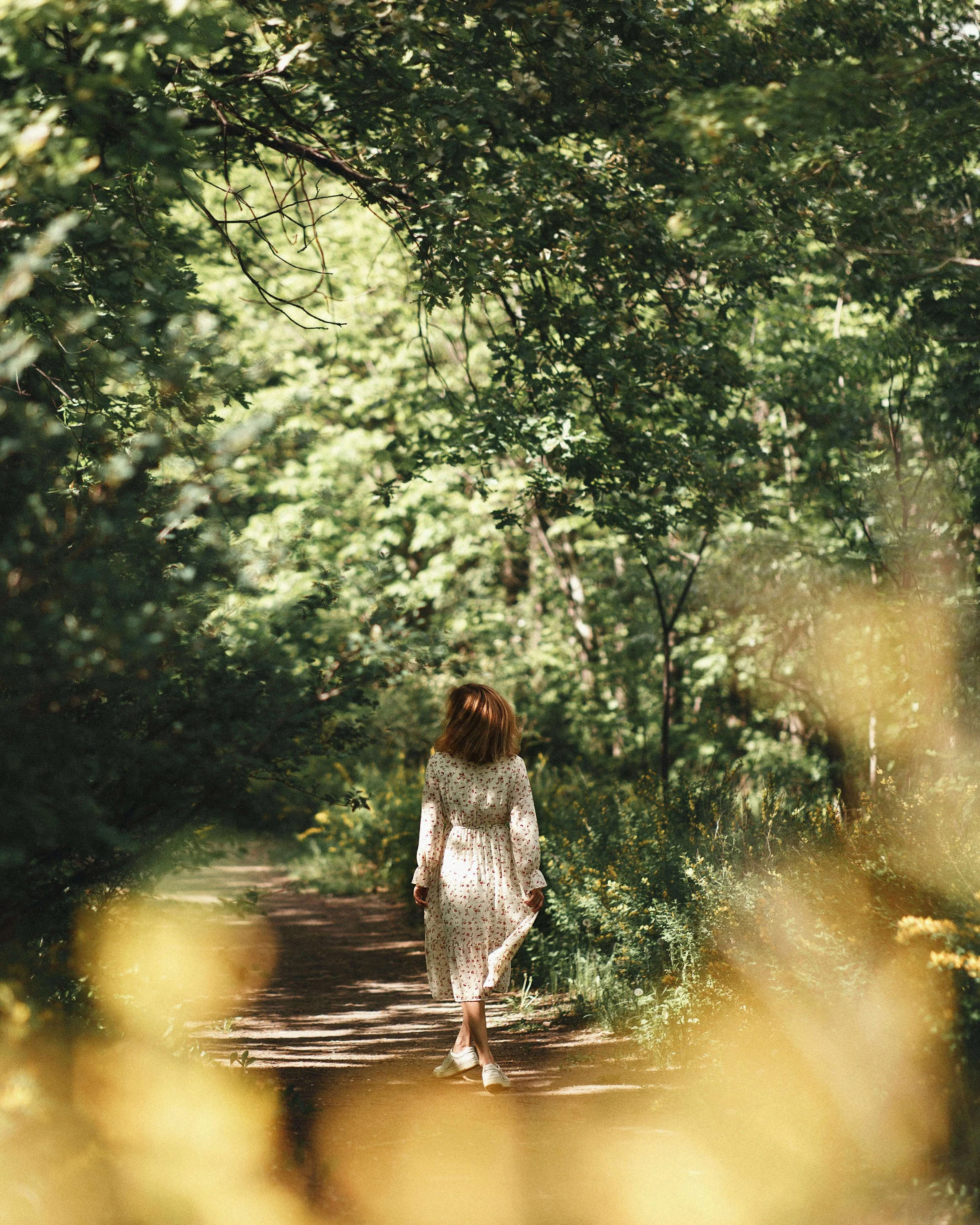 A woman in a white floral dress walking on a forest trail surrounded by green trees and sunlight filtering through the leaves.