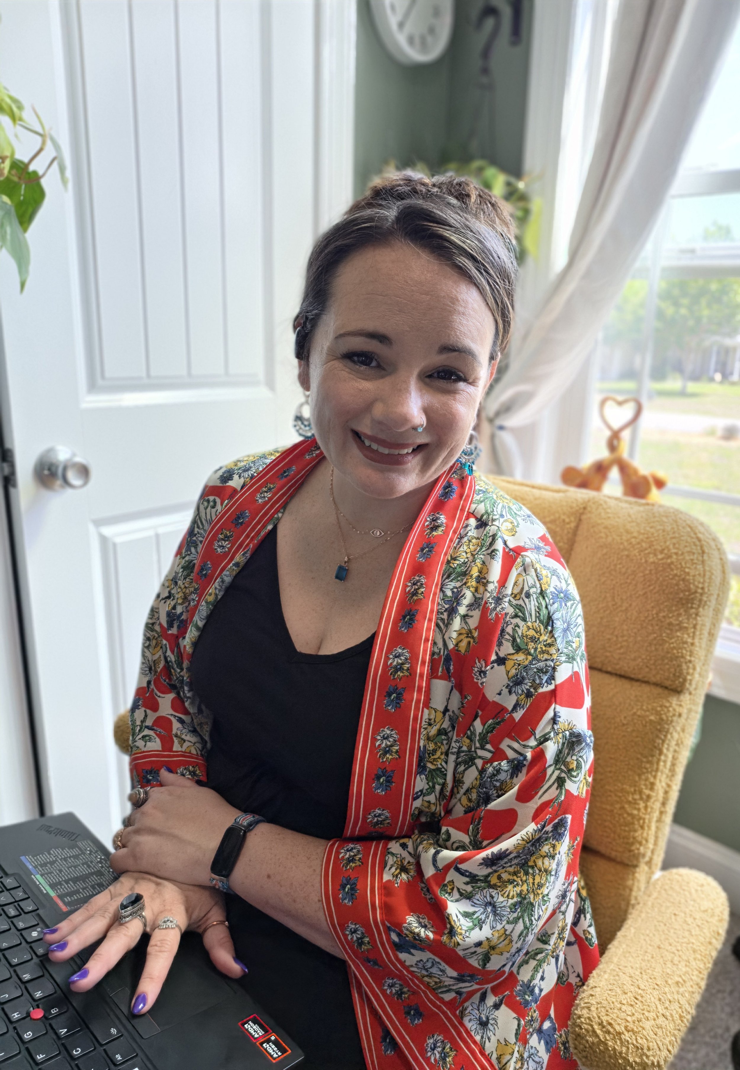 A smiling woman with short brown hair, wearing a black shirt and a colorful floral robe, sitting in a yellow armchair near a window, with a laptop on her lap, in a cozy room with light green walls and a white door.