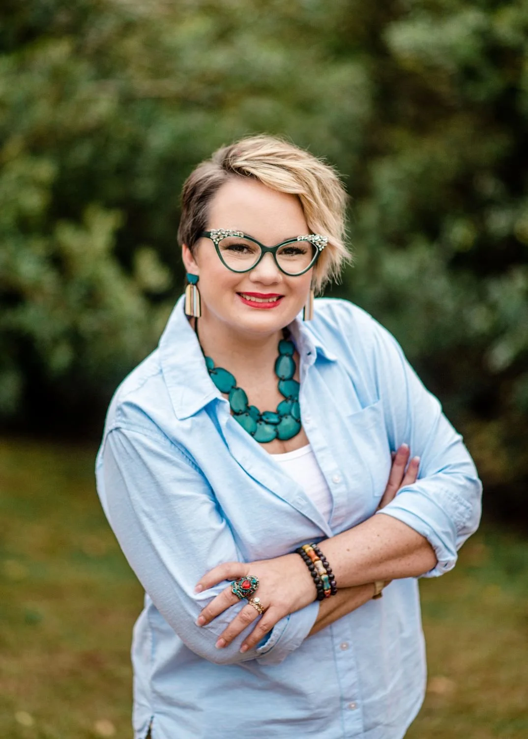 Woman with short blonde hair wearing glasses with embellishments, a blue necklace, earrings, and a light blue shirt standing outdoors with arms crossed.