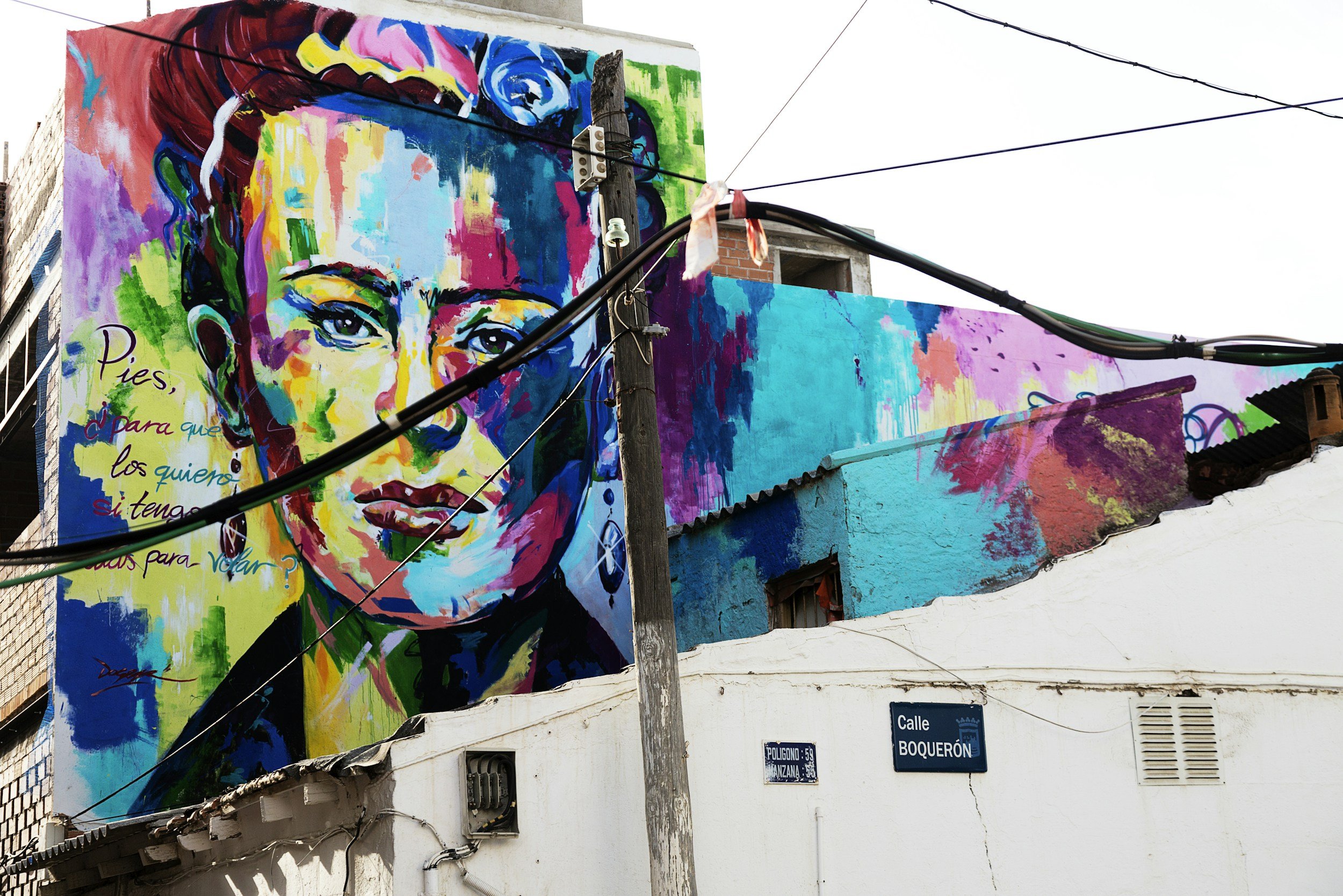 Colorful mural of a woman's face on a building, featuring vibrant blues, yellows, and pinks, located above a street sign reading 'Calle Boquerón.'