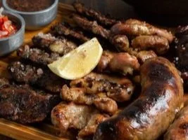 Grilled meat platter with assorted sausages and ribs, garnished with a lemon wedge, and accompanied by small bowls of sauce.