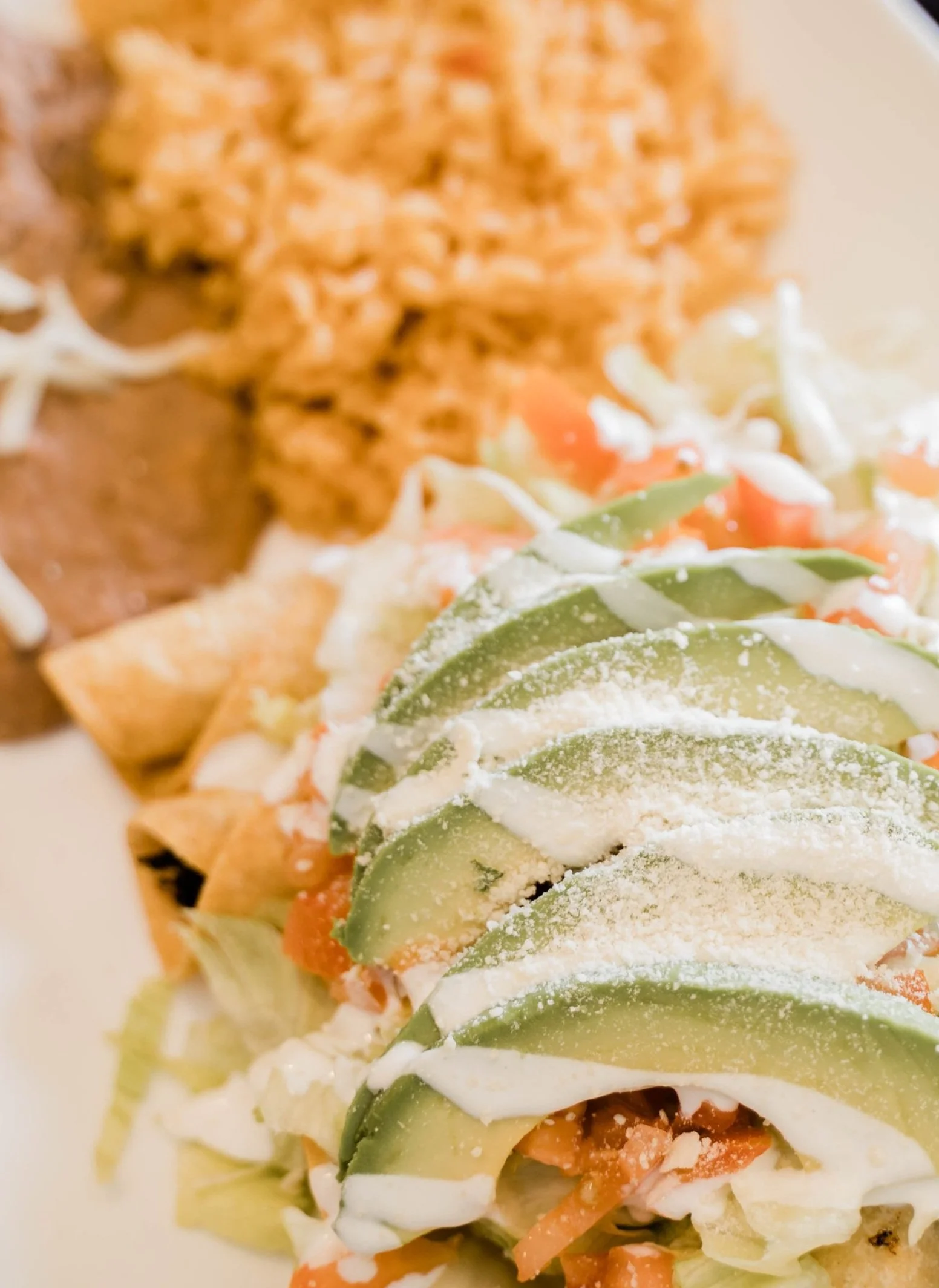 Mexican dish with avocado slices, lettuce, tomatoes, sour cream, and cheese, served with refried beans and rice on a plate.
