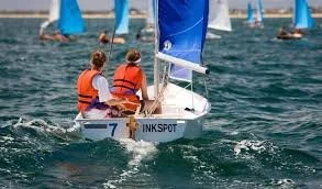 Two children in orange life jackets sailing a small boat on Albert Park lake with blue sails.