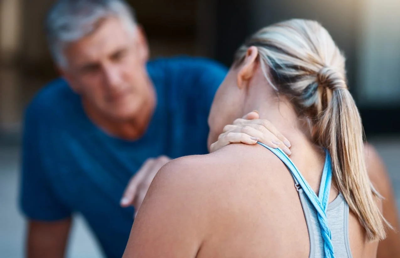 A woman in athletic clothing holds her neck in pain while a man in a blue shirt provides support and comfort.